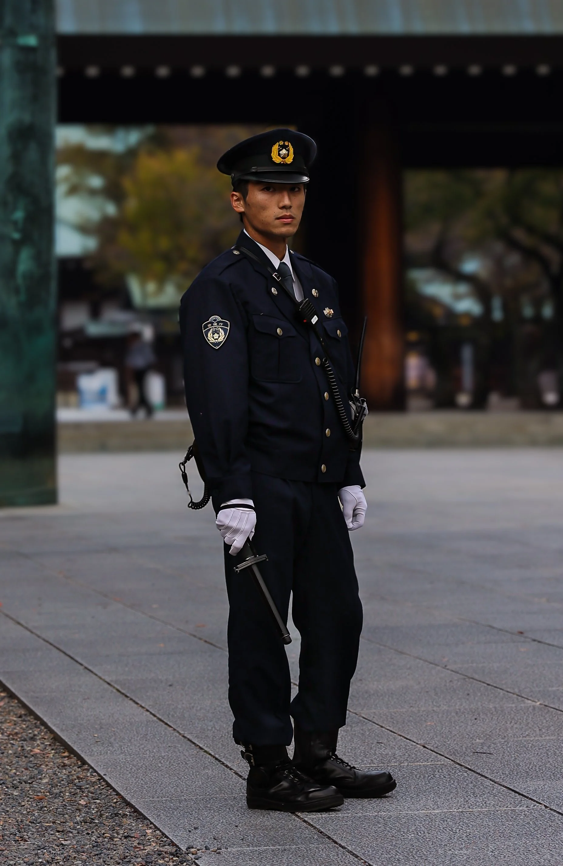 A police officer in uniform standing outdoors, holding a baton, with a serious expression, under a bridge or overpass, with trees and a blurred background.