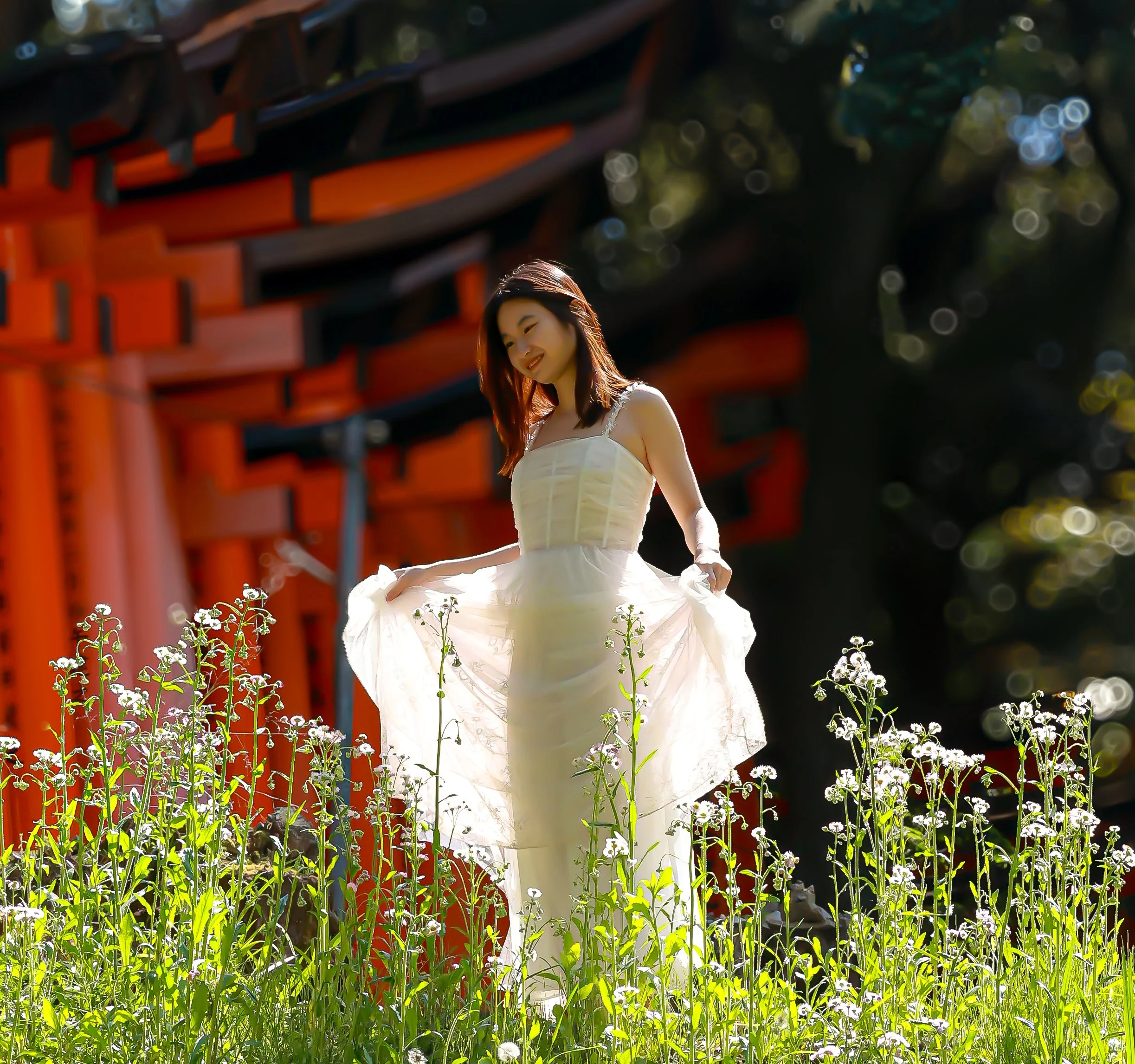 A young woman in a white dress is standing outdoors among small white flowers, with a large red structure in the background and trees with sunlight filtering through.