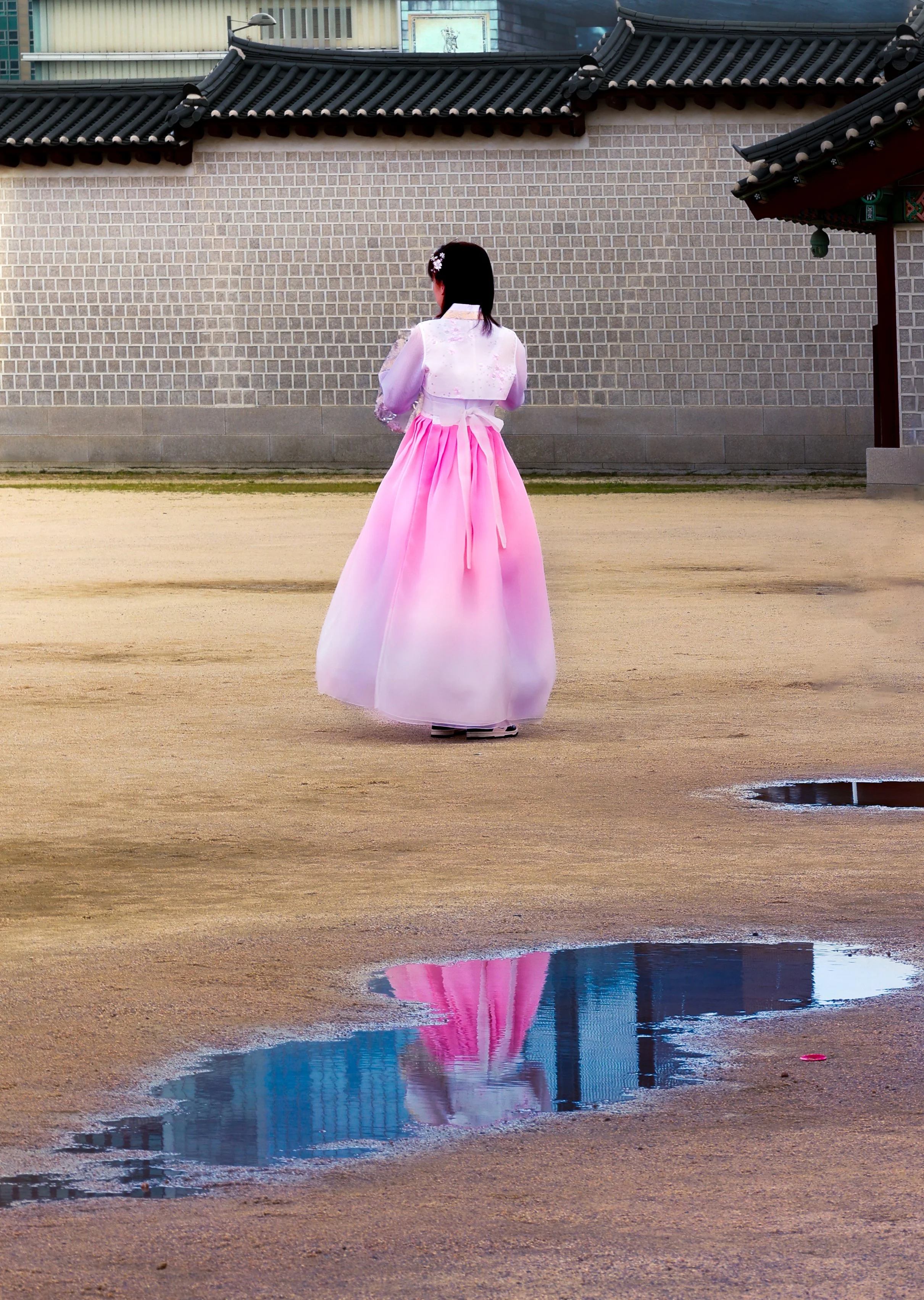 A woman dressed in a traditional pink and white hanbok standing on a dirt ground, with a wall and traditional Korean roof behind her. Her reflection is visible in a small puddle of water on the ground.