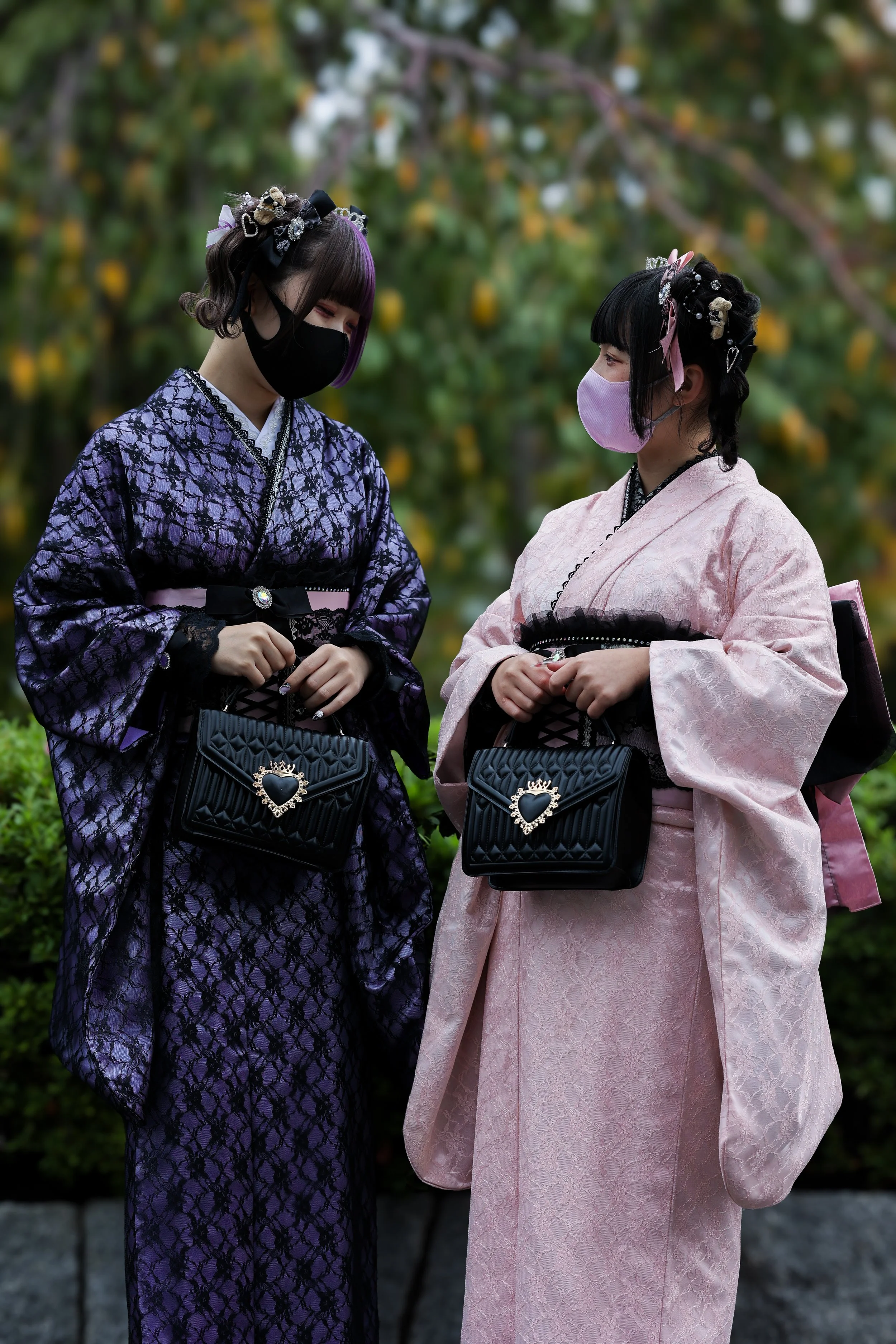 Two young women dressed in traditional Japanese kimonos and wearing face masks, standing outdoors in front of a blurred background of green foliage.