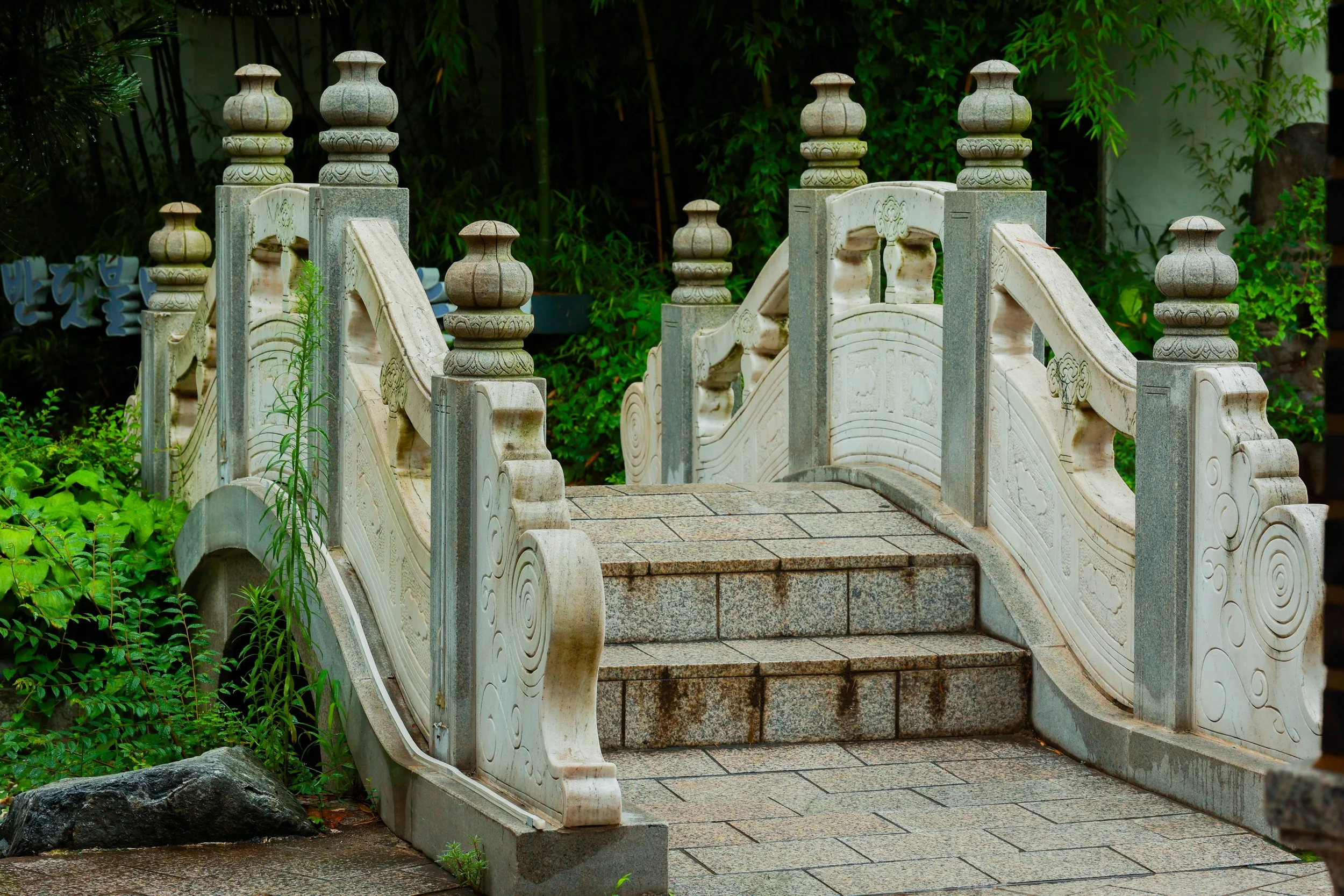 A small stone bridge with carved railings and pillars, surrounded by lush green plants and trees.