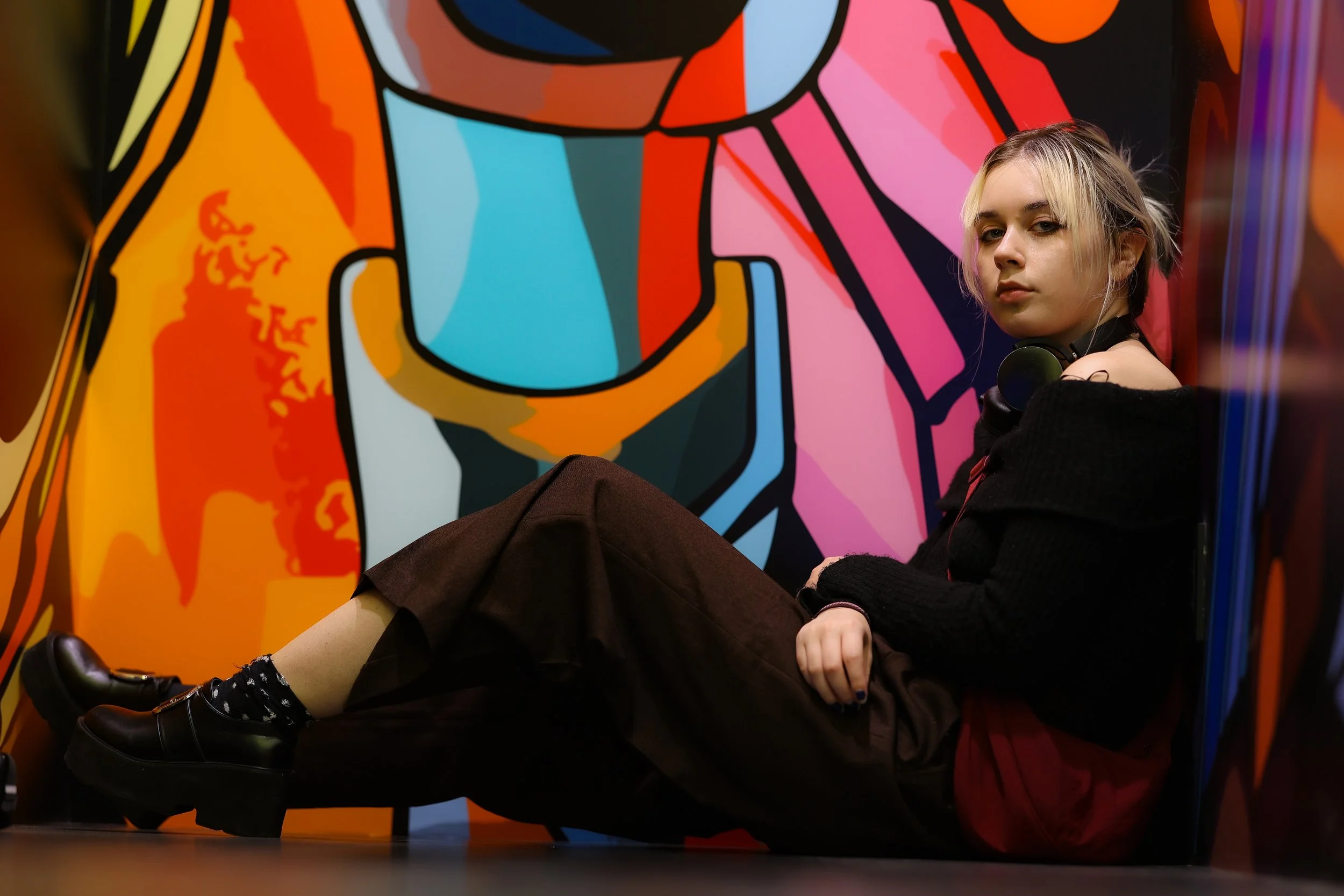 A young woman with blonde hair and a black off-shoulder top sitting on the floor against a colorful, abstract mural with bold lines and bright colors.