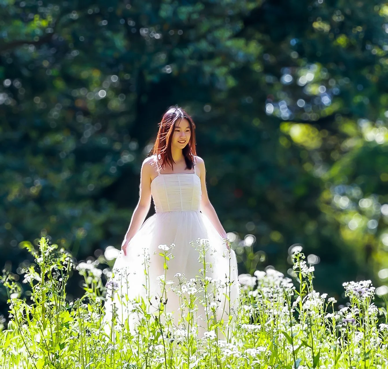 A young woman in a white dress standing in a field of flowers with a blurred green forest background.