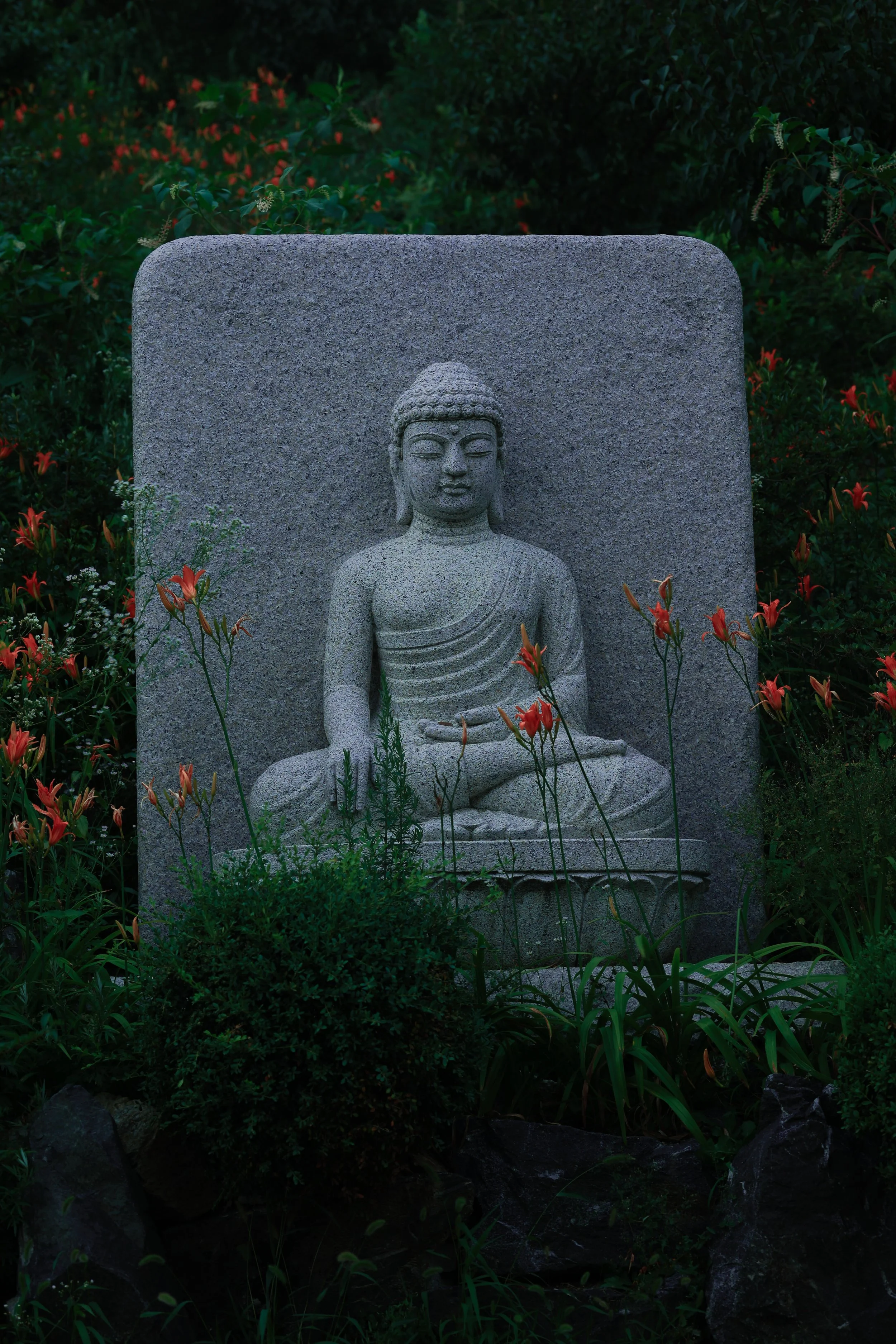 A stone sculpture of a seated Buddha with closed eyes, set against a dark background with greenery and flowers.