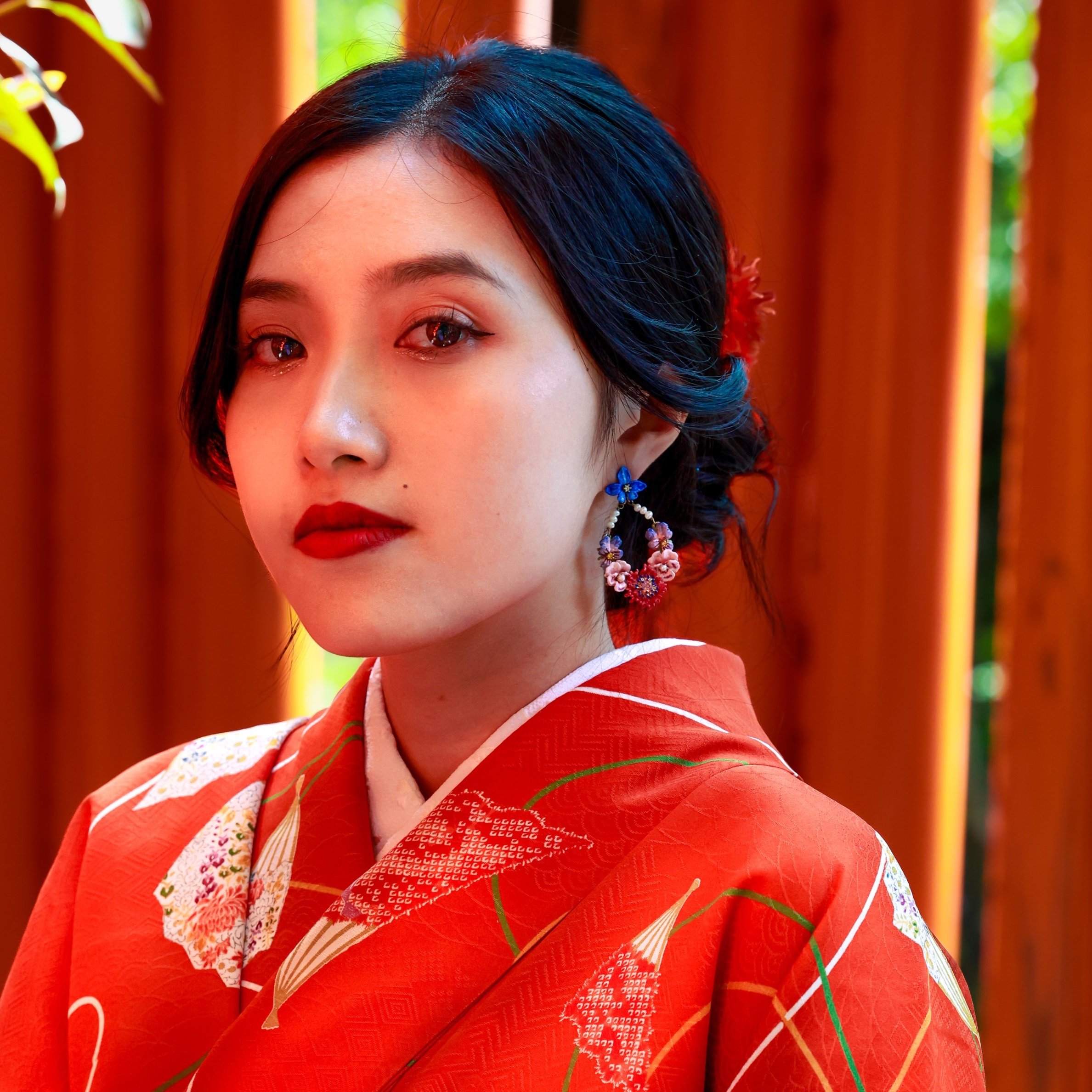 Close-up of a woman wearing a traditional red kimono with floral and geometric patterns, earrings, and makeup.