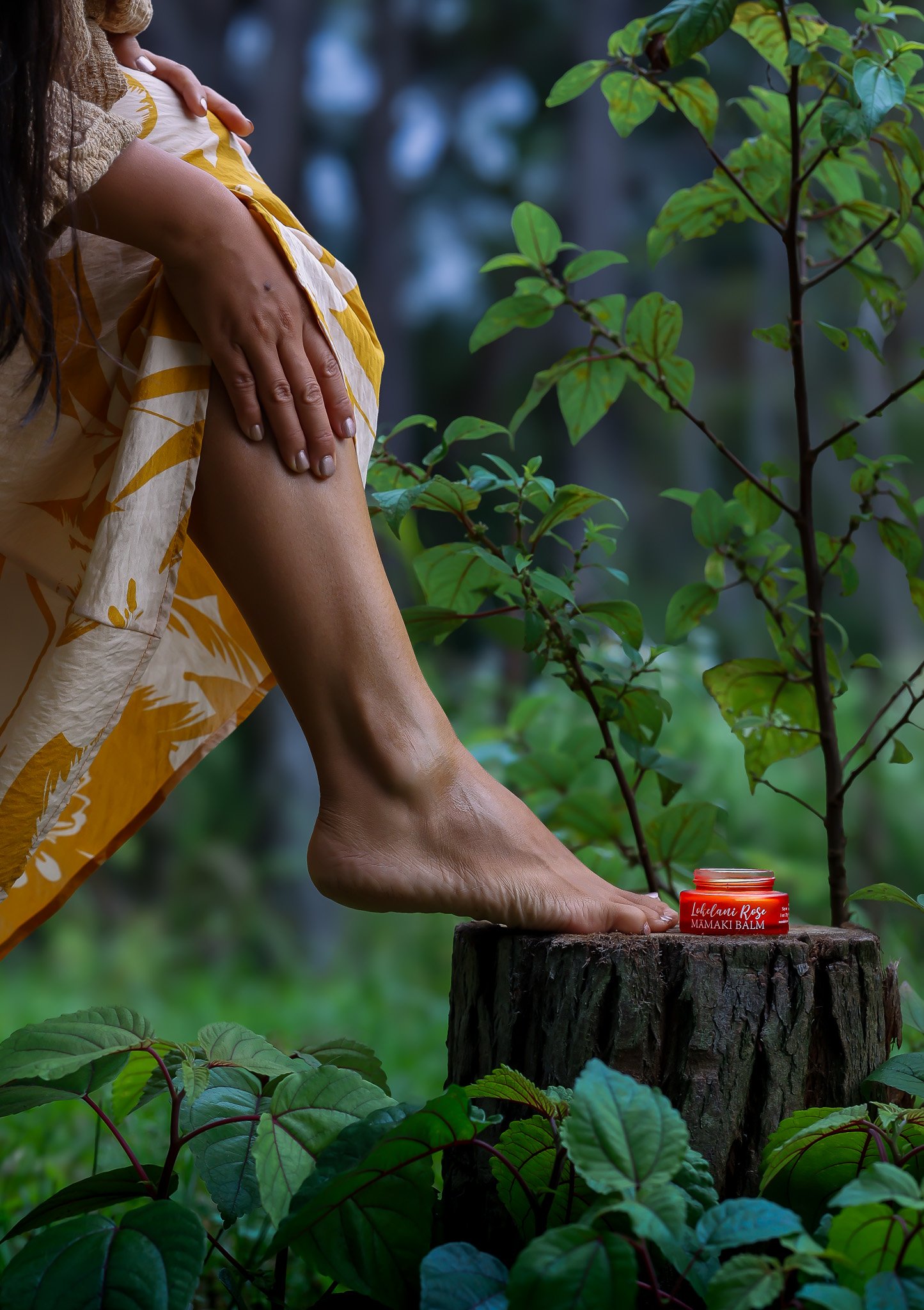 A person in yellow and white clothing kneels on a tree stump outdoors, with one hand on their knee and the other resting on a small jar of balm labeled 'Lululani Rose Mama Balm.' The scene is surrounded by green foliage and plants, suggesting a natural setting.