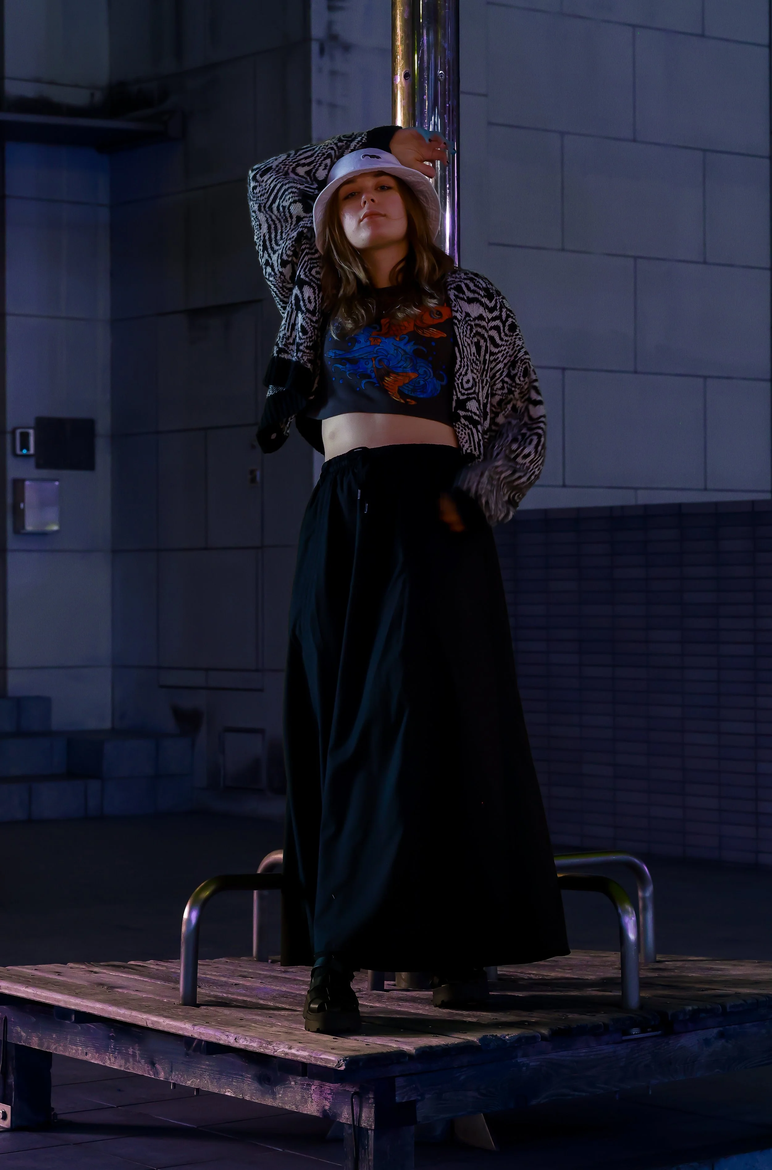 A young woman standing on a wooden platform outdoors, wearing a white bucket hat, a cropped graphic t-shirt, a patterned jacket, and loose dark pants, with one hand resting on her forehead and the other in her pocket, illuminated by dim artificial li