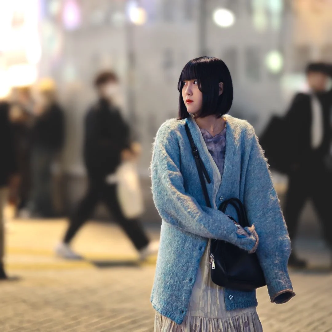 A woman with black bob haircut and bangs stands on a city street at night, wearing a grey cardigan and holding a black bag, with blurred pedestrians walking in the background.