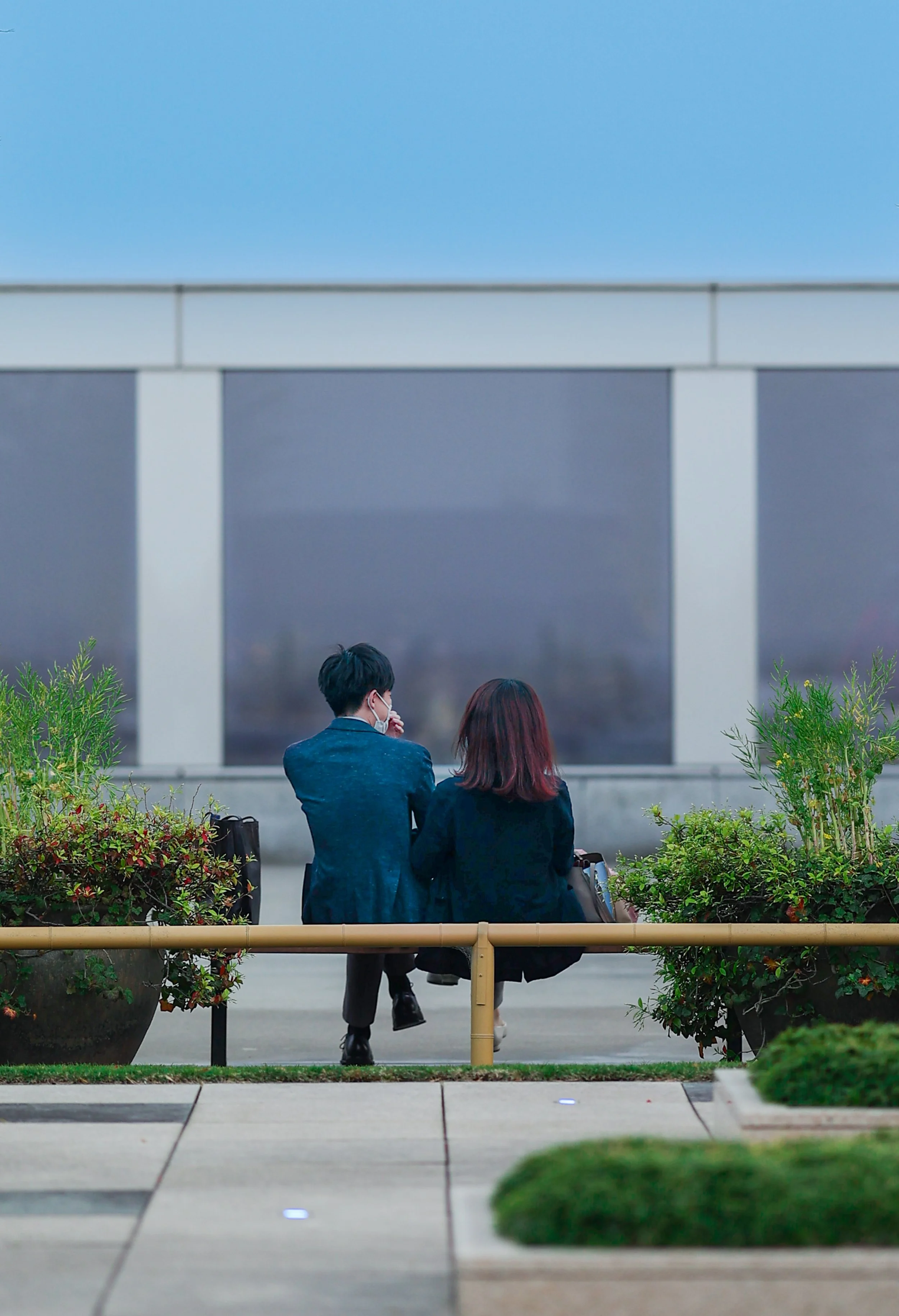 Two people sitting on a bench outside, facing a building. One person wears a mask, and they are surrounded by greenery.