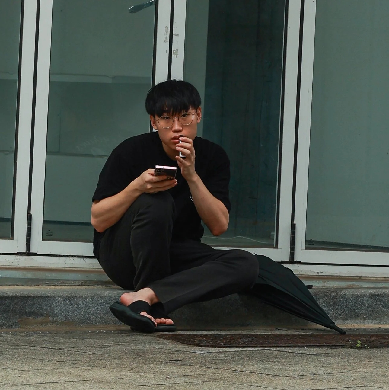 A young man with glasses, black hair, wearing a black t-shirt and black pants, sitting cross-legged on a sidewalk near glass-door windows, holding a smartphone in his right hand and a cigarette in his left hand, with a black umbrella resting on the g