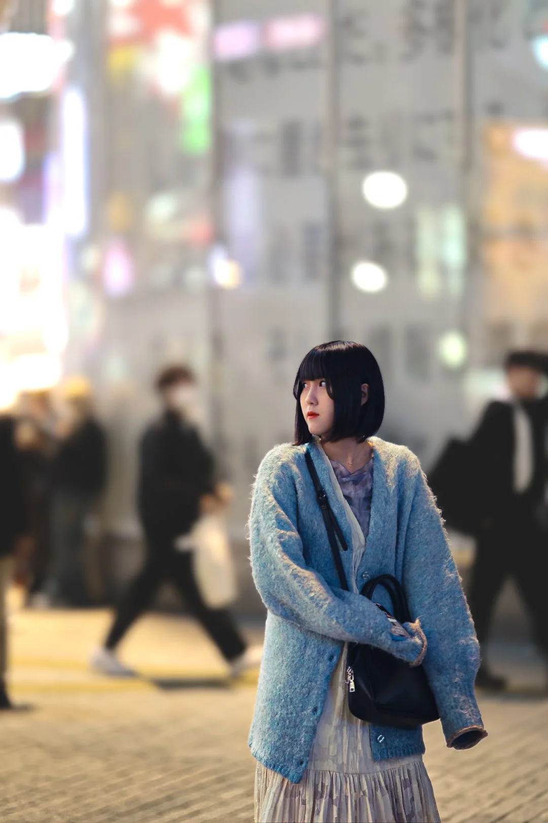 A young woman with black hair and bangs stands on a city street at night, wearing a blue cardigan over a dress, with her arms crossed, and is surrounded by blurred people walking in the background.