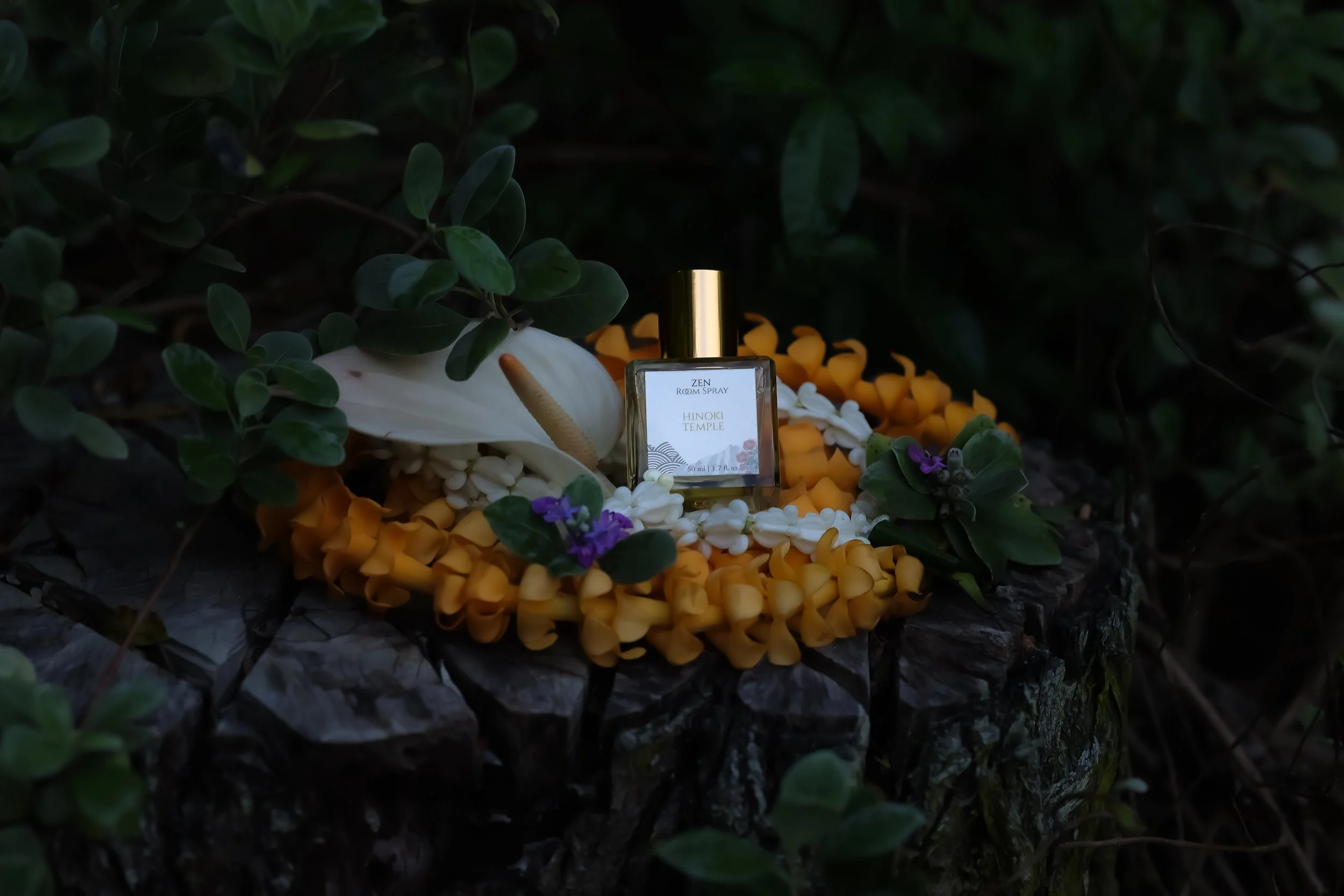 A perfume bottle labeled Zen Room Spray placed on a bed of yellow, white, and purple flowers, surrounded by green foliage with a dark background.