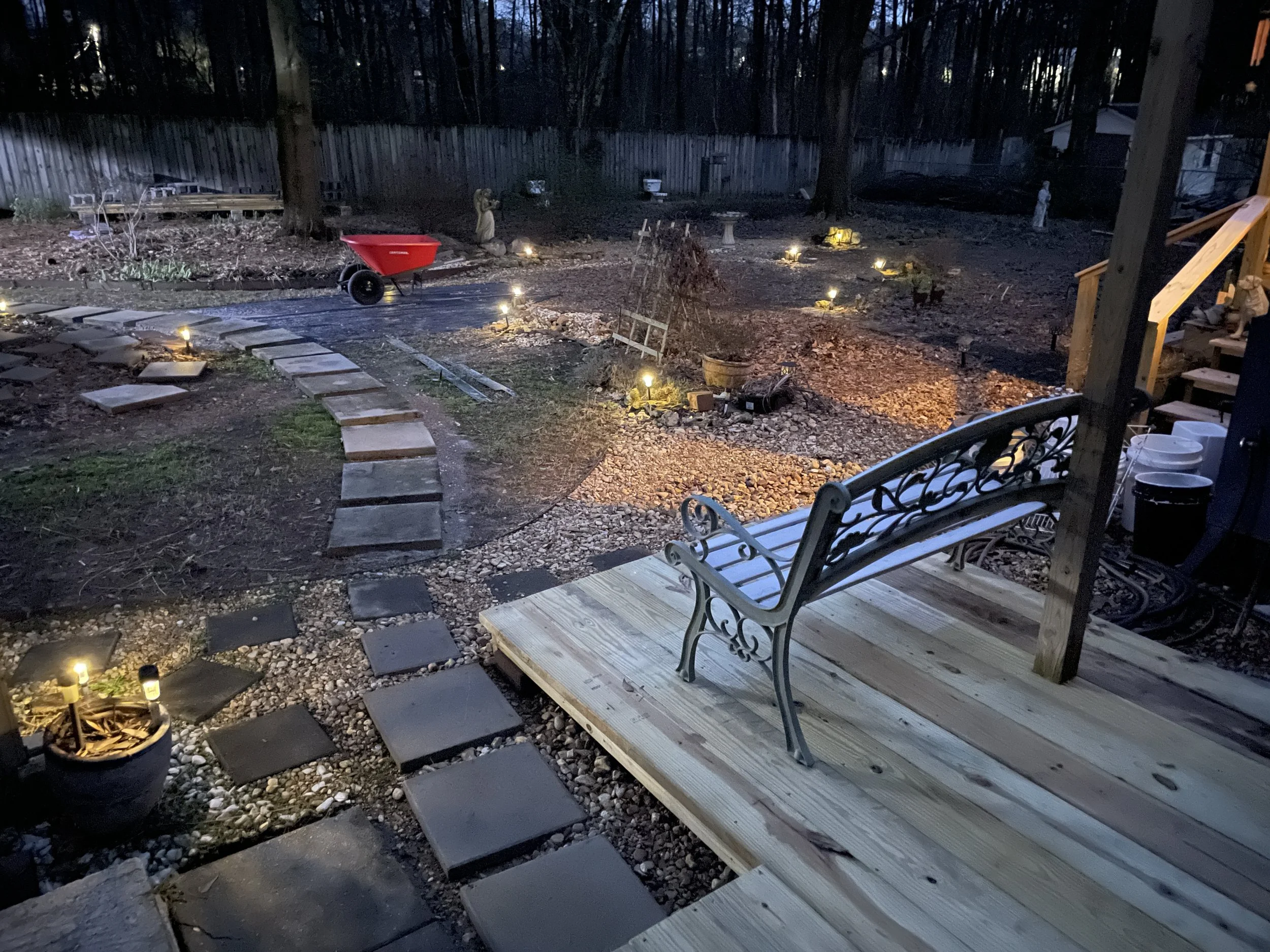 A backyard at dusk with a wooden deck and benches, paved pathways, a red wheelbarrow, and outdoor lighting.