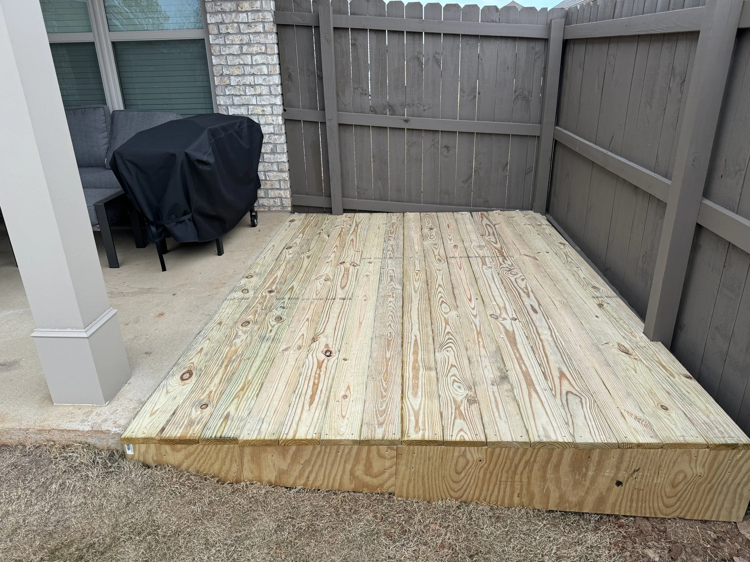 New wooden deck platform adjacent to a covered outdoor grill area, with a wooden privacy fence and patio furniture in the background.