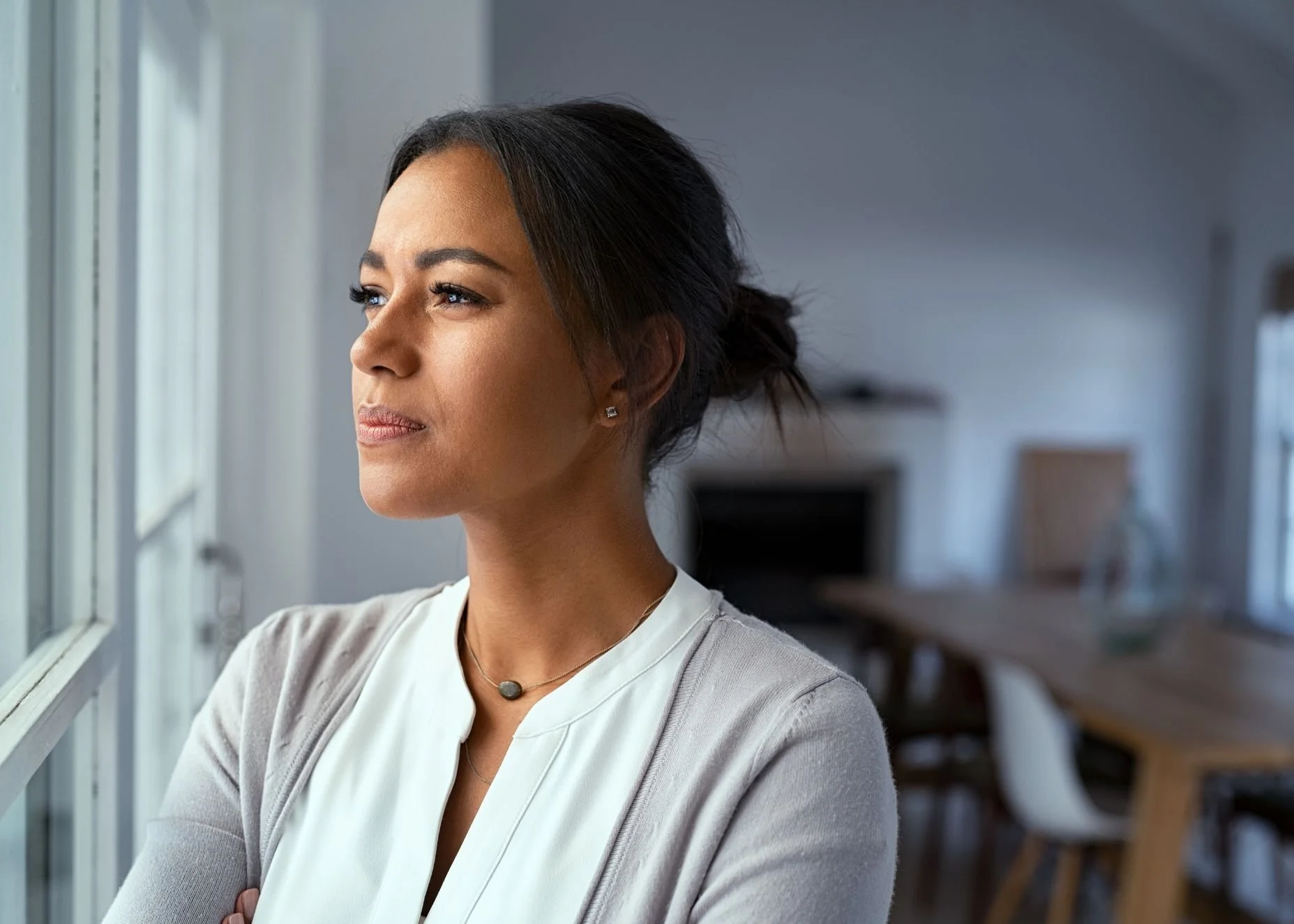 a young woman stands indoors looking out a window with a contemplative expression