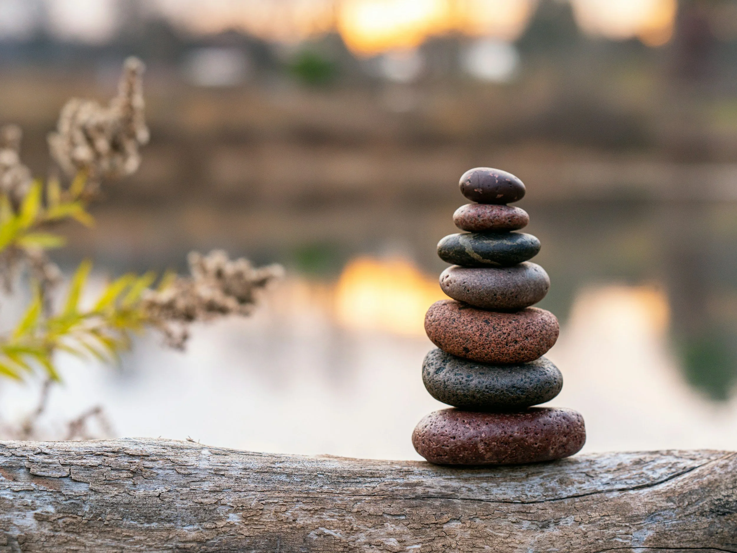 A stack of seven smooth, rounded stones balanced on top of each other on a weathered wooden log. The background features an out-of-focus sunset or sunrise scene with warm tones and blurred landscape elements.