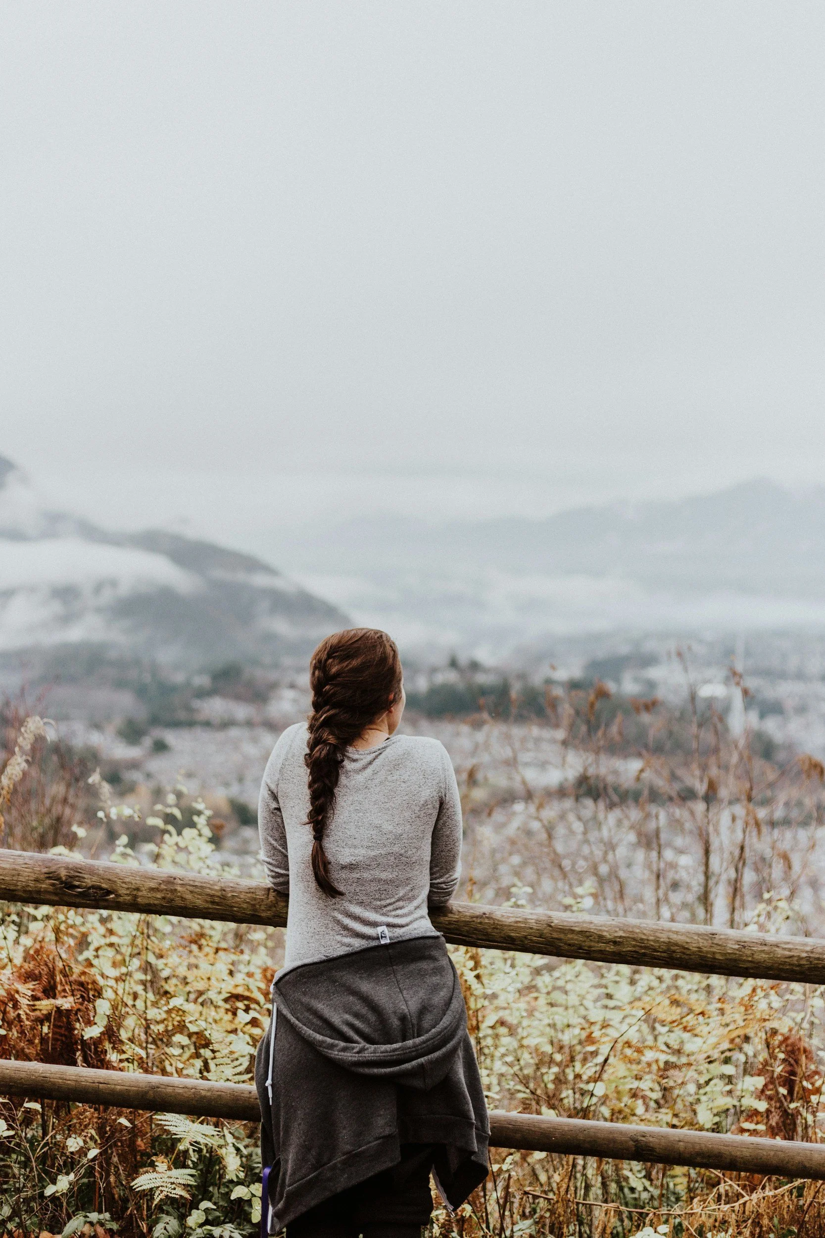 a young woman with hair braided and a jacket around her waist stands near a wooden split rail fence with back turned overlooking an out of focus landscape with a dusting of snow