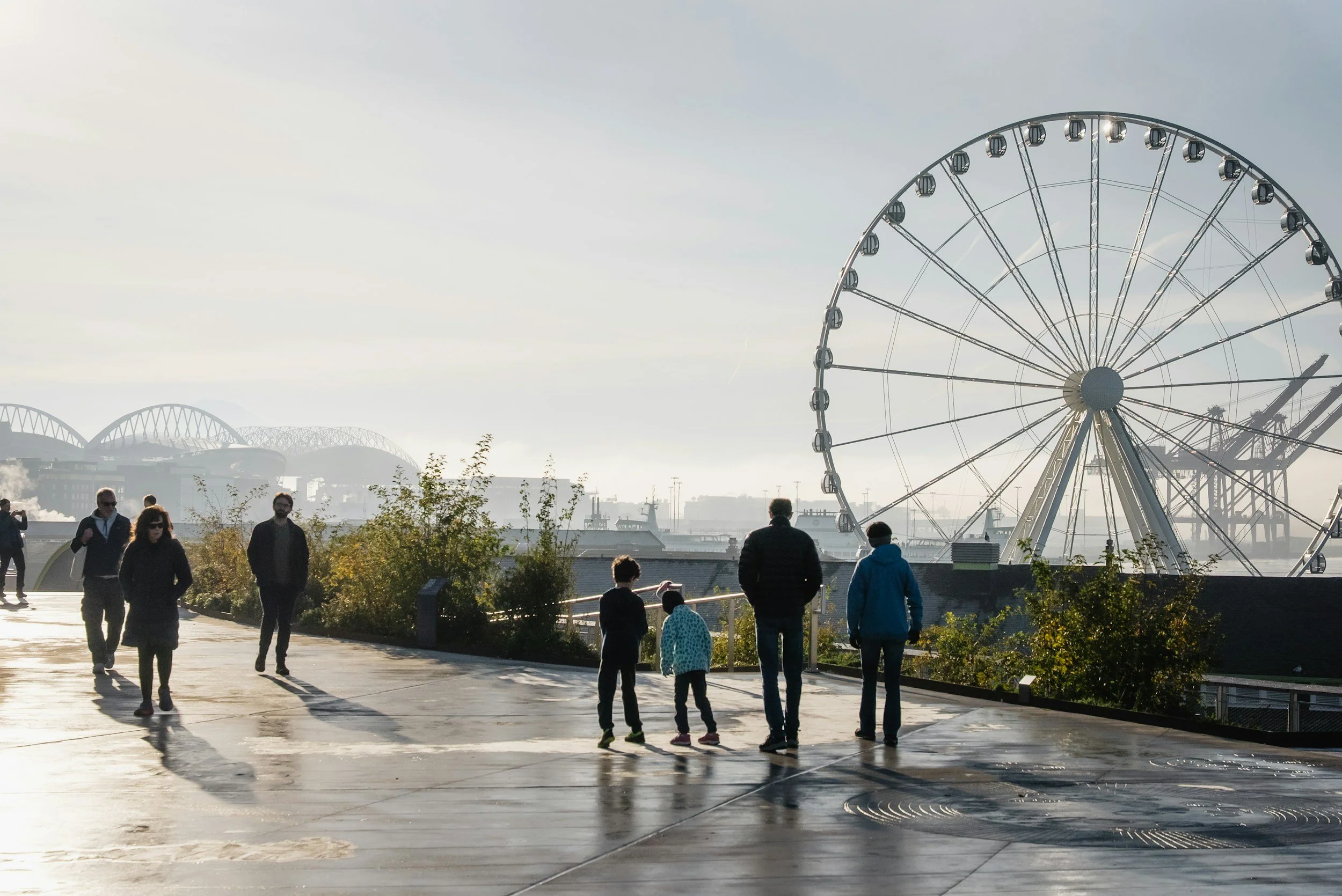 People walking along a promenade with the Seattle Great Wheel in the background, near a harbor with ships and a bridge.