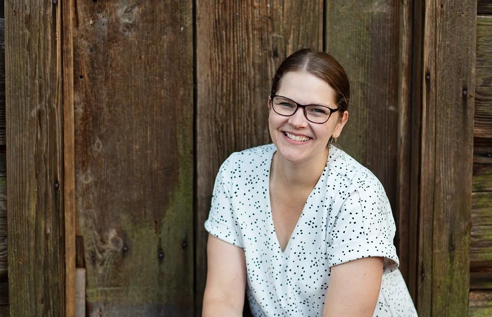 A smiling woman with brown hair, glasses, and a white shirt with black dots sitting outdoors against a wooden fence.