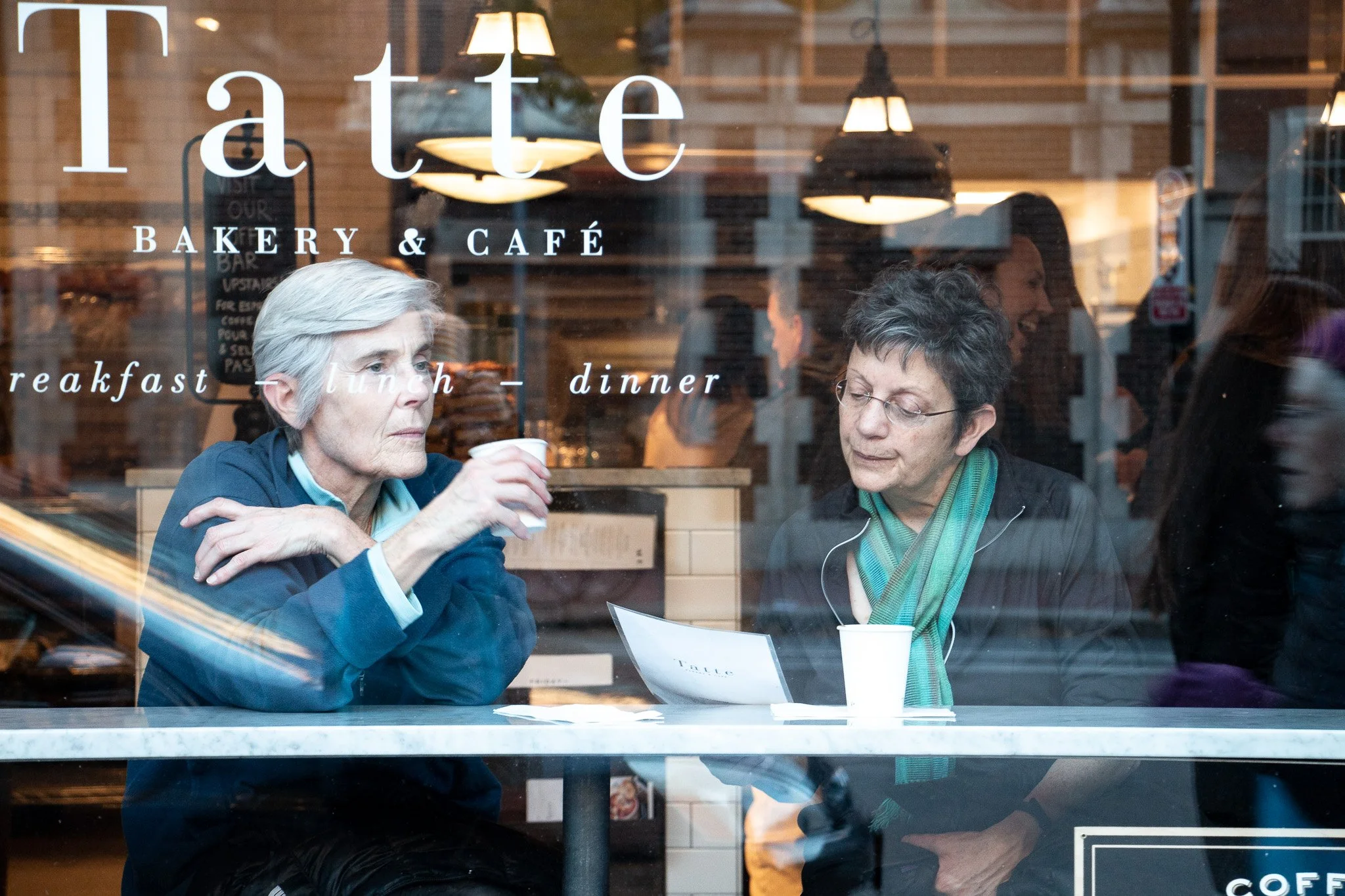 Two elderly women sitting behind a window at a bakery and cafe, having a drink and looking at a piece of paper, with the shop's name and menu items displayed on the glass. Photo by Hannah Loomis