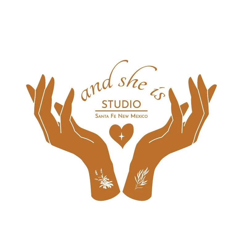 Logo featuring two brown hands with floral tattoos, holding a heart, with the text 'and she is studio Santa Fe New Mexico'.  Design by Hannah Loomis.