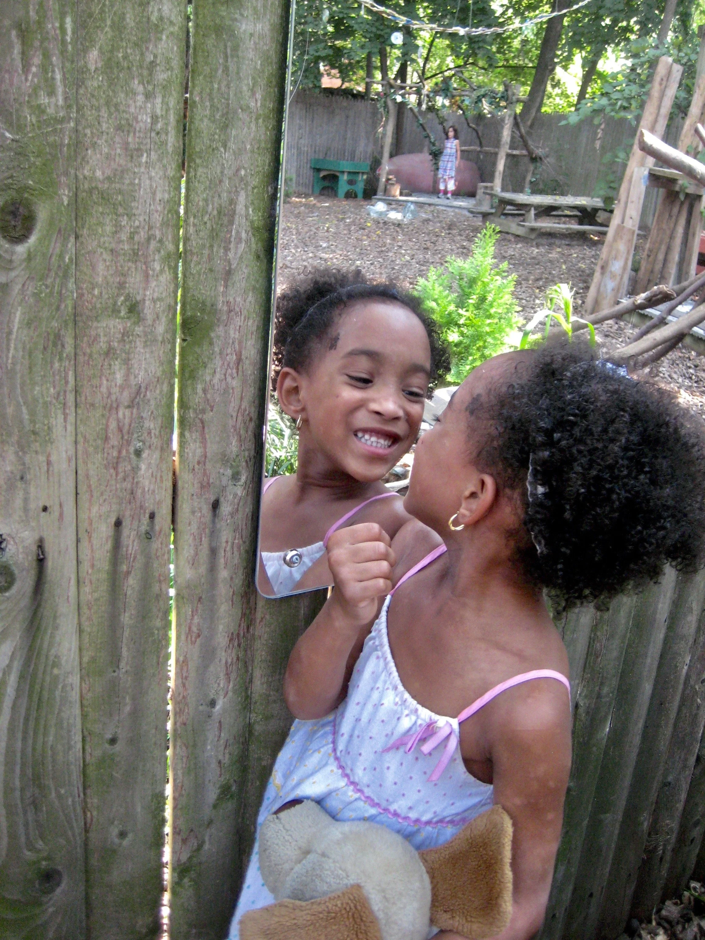 A child smiles at her reflection in an outdoor mirror, supporting self-awareness, social-emotional learning, and nature-based play in an early childhood setting