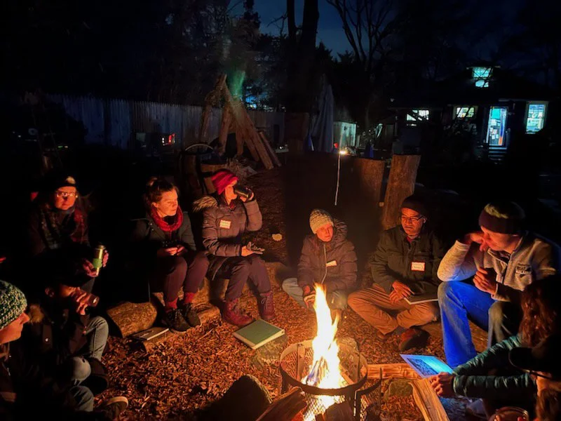 Parents gather around an evening campfire for cooperative nursery school parent education meeting in Takoma Park, MD. Families discuss child development topics and build community connections at outdoor preschool event.