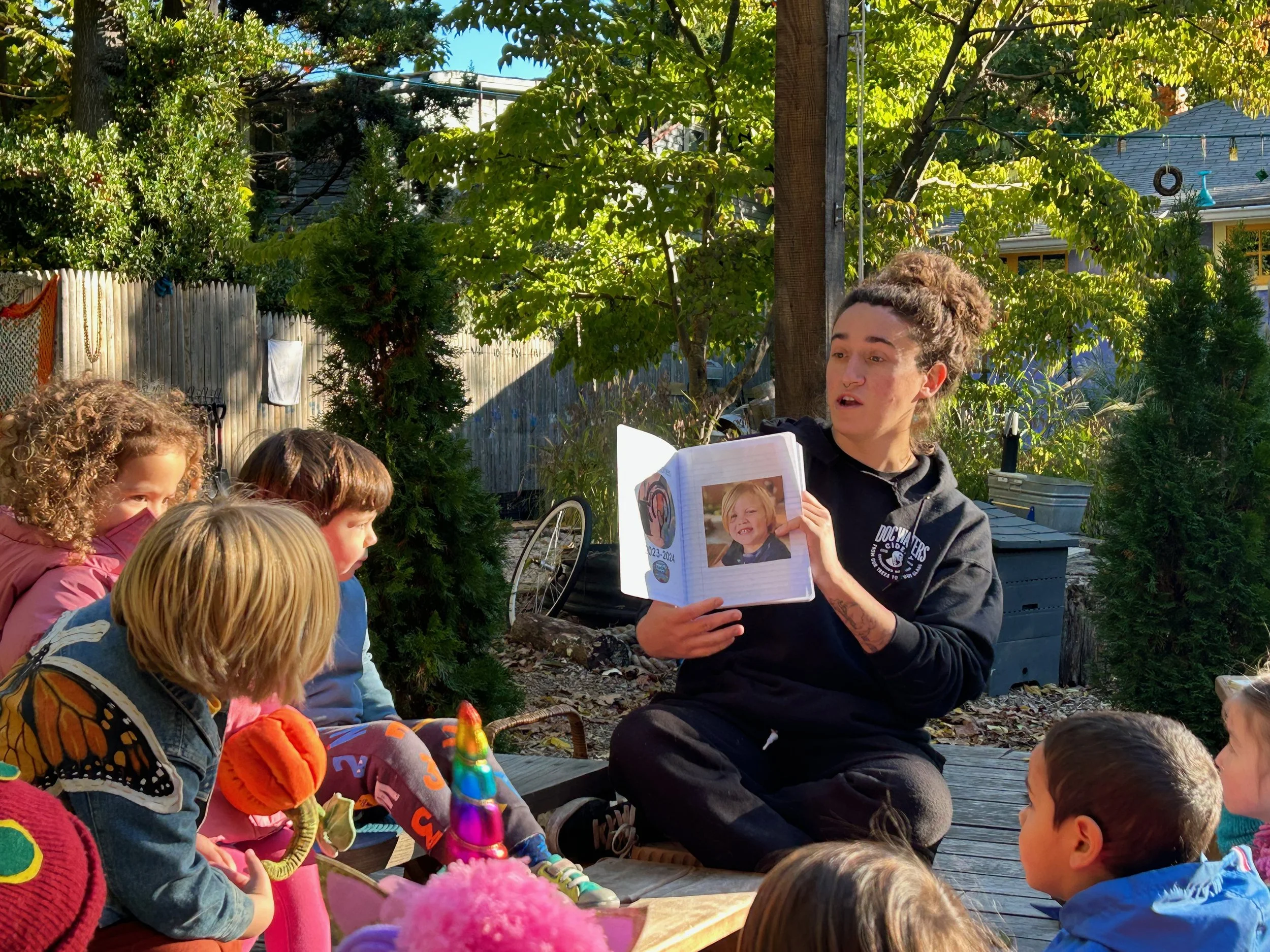 Children gather around the teacher during outdoor story time, immersed in a nature-based learning experience.
