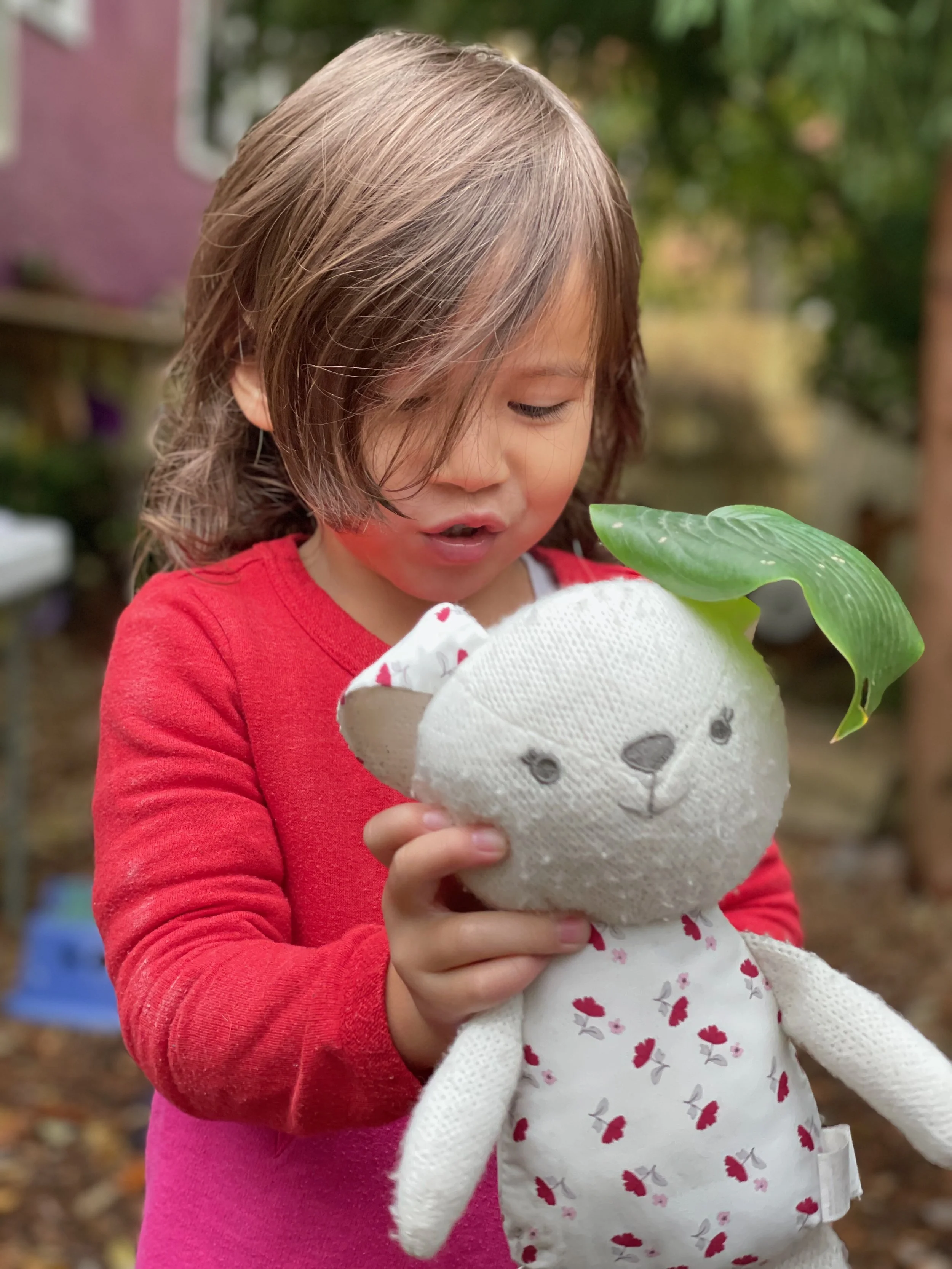 On a rainy day, a child holds a leaf over her stuffed animal—an image of nurturing care, imagination, and the gentle confidence that grows with time.