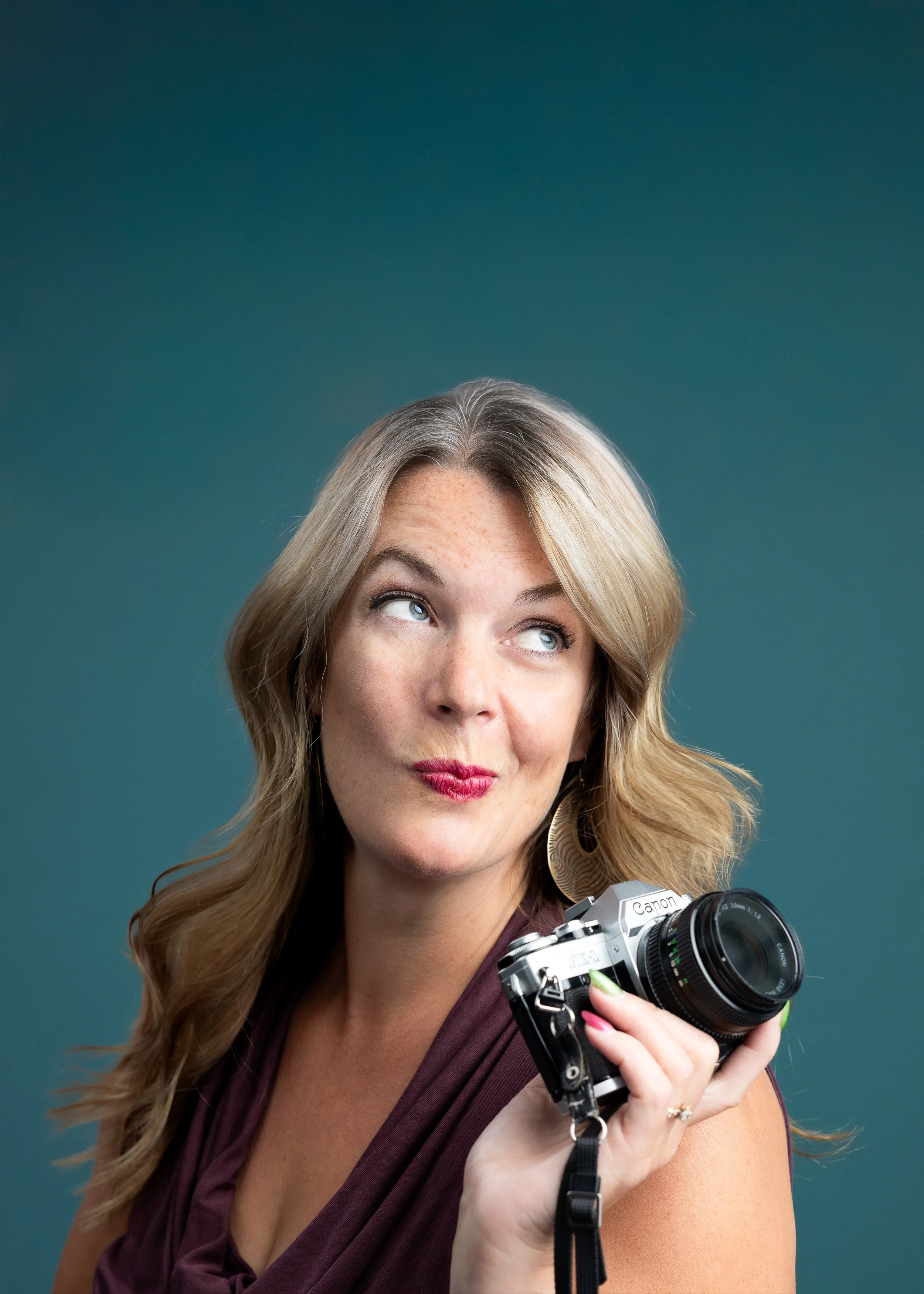A woman with blonde hair and freckles holding a Canon camera, looking upward with a playful expression, wearing a maroon top and large earrings against a teal background.