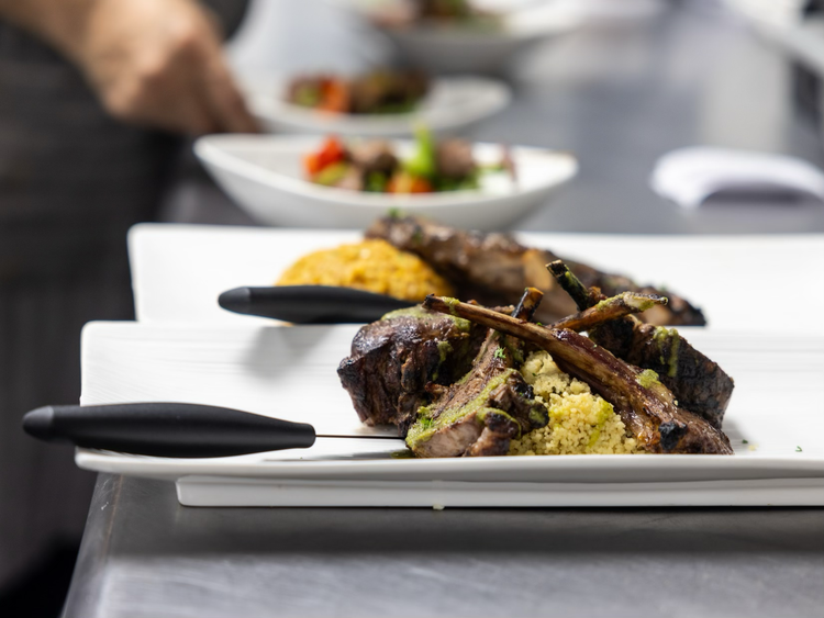 Close-up of a white rectangular plate with grilled lamb chops and mashed potatoes, on a stainless steel serving surface, with a blurred background of other plates and a person's hand.