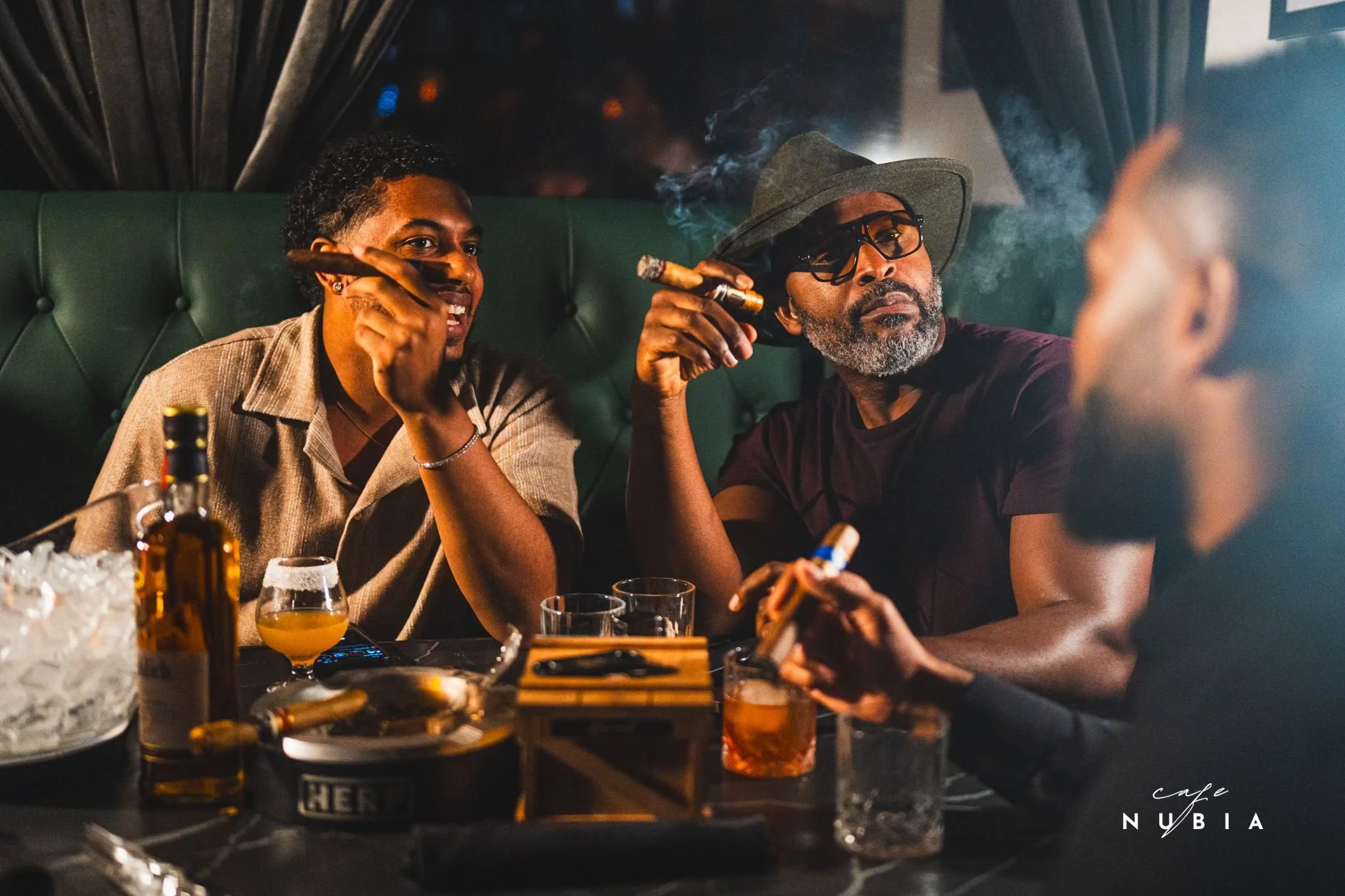 Two men sitting at a bar, smoking cigars, engaging in conversation. The man on the left is smiling, wearing a beige shirt, and the man on the right has a gray beard, glasses, a wide-brim hat, and is looking seriously. There are glasses and bottles on the bar, and the setting appears to be a dimly lit, upscale bar or lounge.