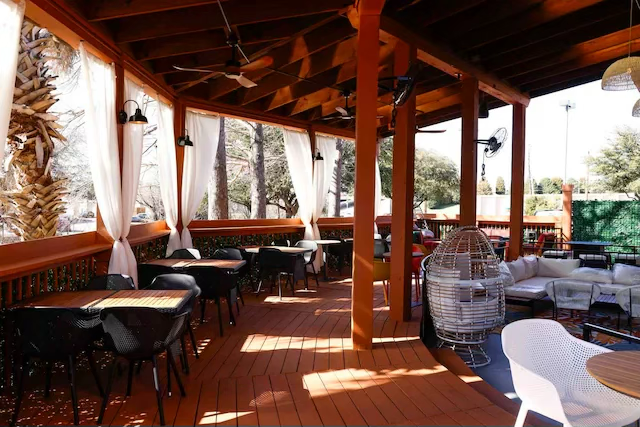 Outdoor patio with wooden floor and ceiling, white curtains, and various seating including tables with chairs and a hanging wicker chair, shaded by trees.