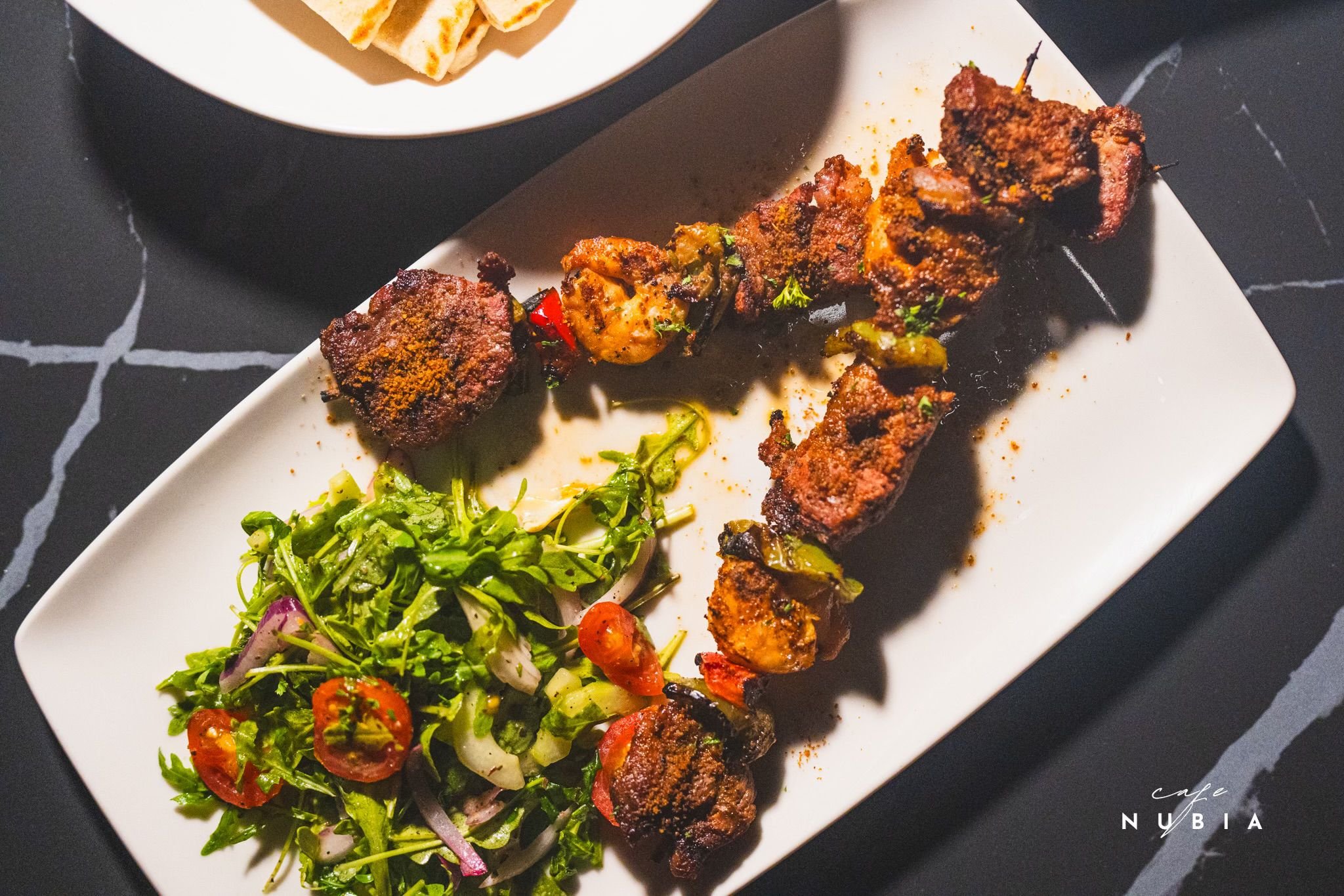 A white rectangular plate with grilled meat skewers, a side salad with cherry tomatoes, red onions, and greens, and a small bowl of flatbread pieces. The background is black with white veining.