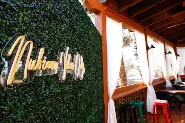 Green hedge wall with yellow neon sign that says 'Mubian House' in a covered outdoor seating area with white curtains and colorful stools