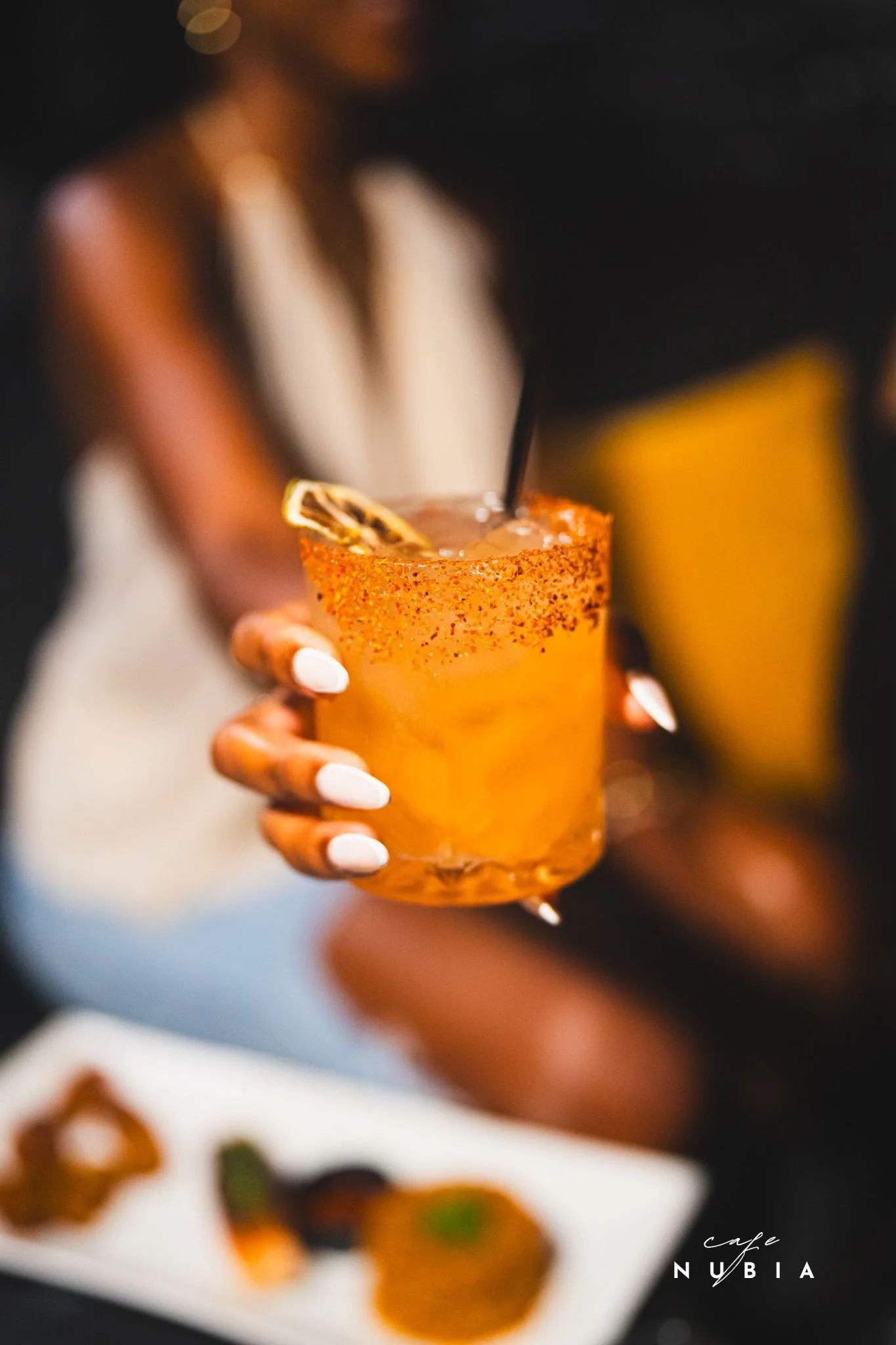 A person holding a drink garnished with a lemon slice, with a blurred background of appetizers on a white plate.