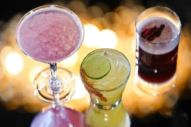 Three colorful cocktails on a reflective surface with blurred lights in the background.