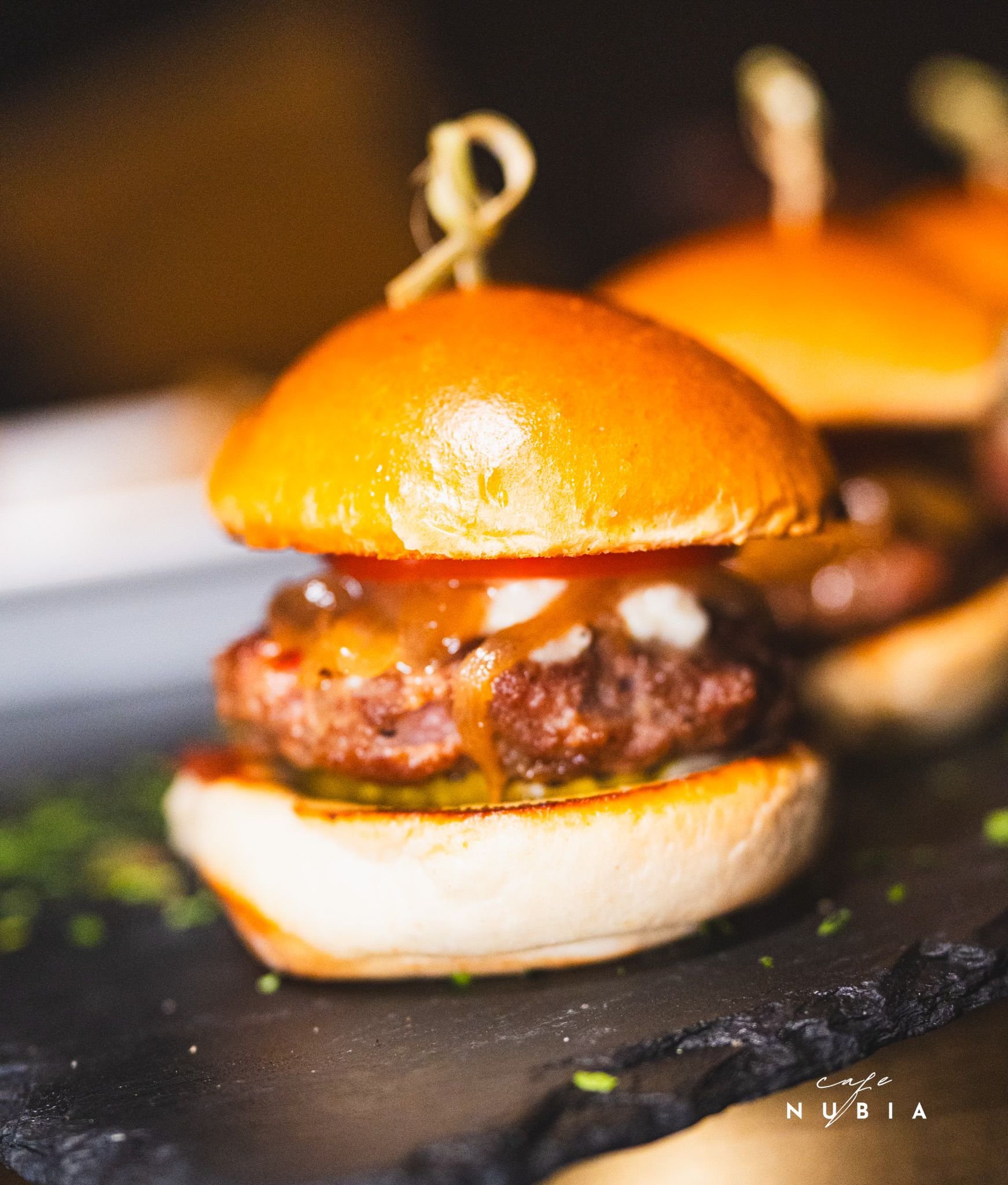 Close-up of a burger with a toasted bun, beef patty, cheese, and toppings, on a black grill or griddle with small green herbs or chives around it.