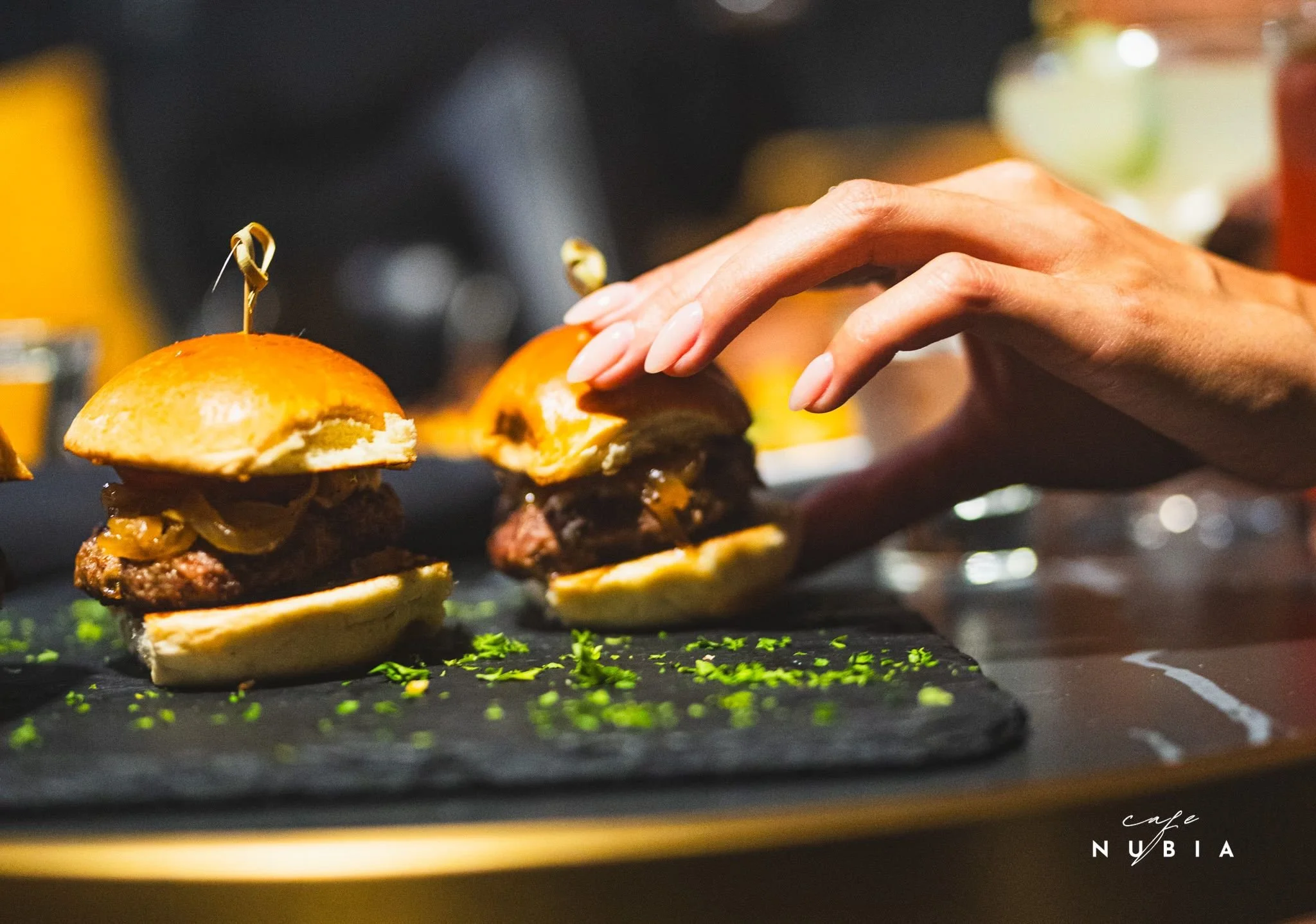 Close-up of a person's hand placing a bun with a meat patty, cheese, and toppings on a skewered burger stack, with another similar burger in the background on a black surface with green herbs garnished around.