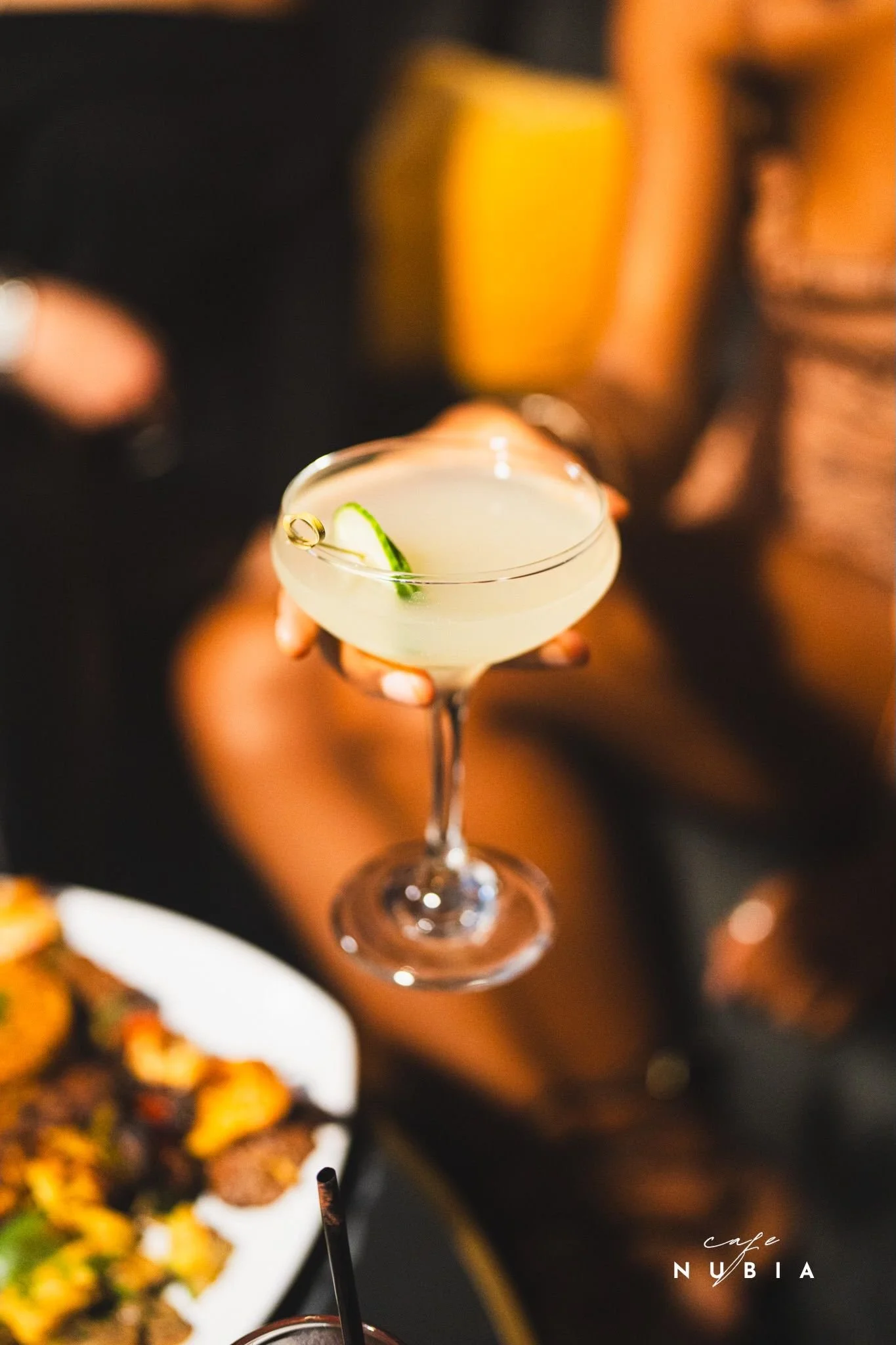 A close-up of a cocktail glass with a lime wedge garnish, held by a person's hand. The background features blurred warm lighting and a hint of a yellow drink, with part of a plate of food visible in the foreground. The text "cafe NUBIA" is located at