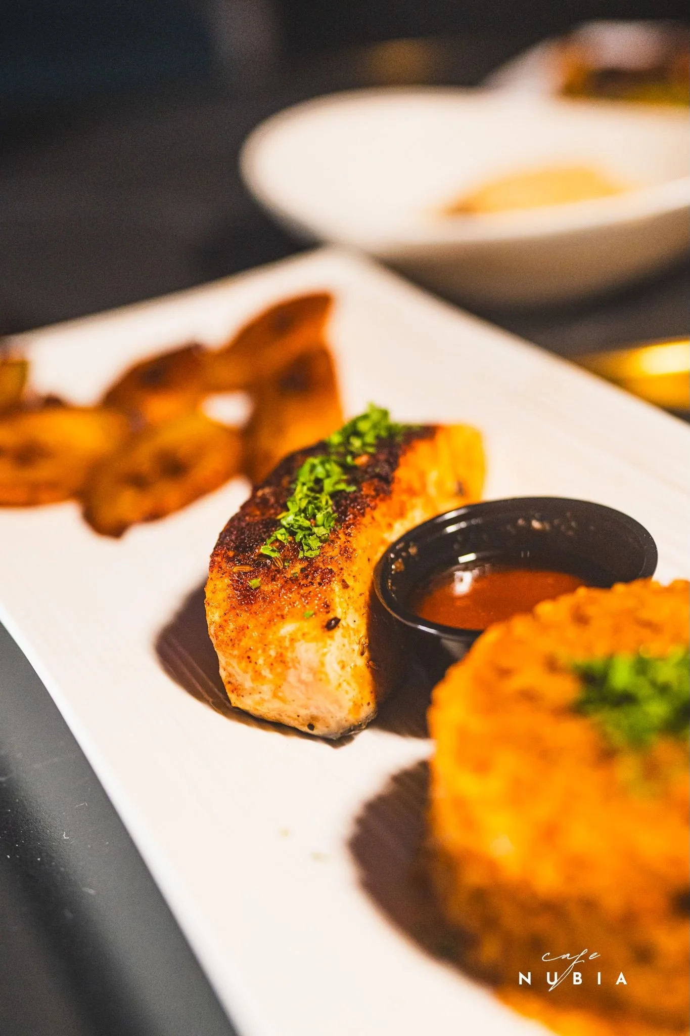 Close-up of a grilled salmon fillet topped with chopped herbs, served on a white plate with a side of dipping sauce. In the background, slices of grilled bacon and a bowl of white sauce are visible. The image has a dark, blurred background and the text "cafe NUBIA" at the bottom right corner.