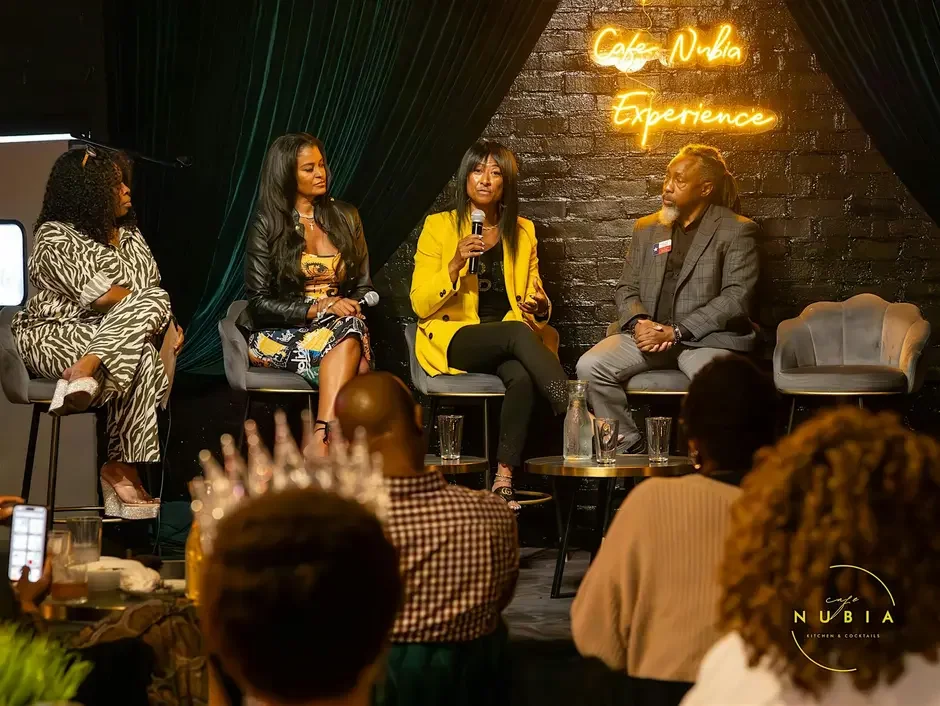 Four people seated on a stage with a black brick wall background, illuminated by a neon sign reading 'Café Nubia Experience,' participating in a panel discussion. The audience is visible in the foreground.