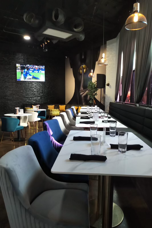 A modern restaurant interior with a long white table set for dining, including glasses and black napkins, with colorful chairs and a wall-mounted TV showing a sports game.