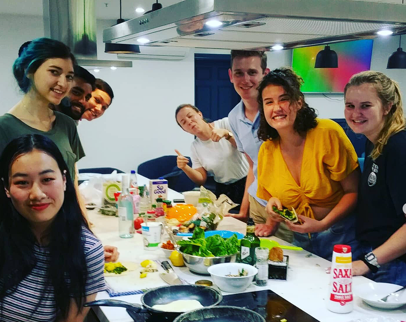 Group of smiling young adults gathered around a kitchen table with food ingredients and cooking utensils, enjoying a cooking workshop.
