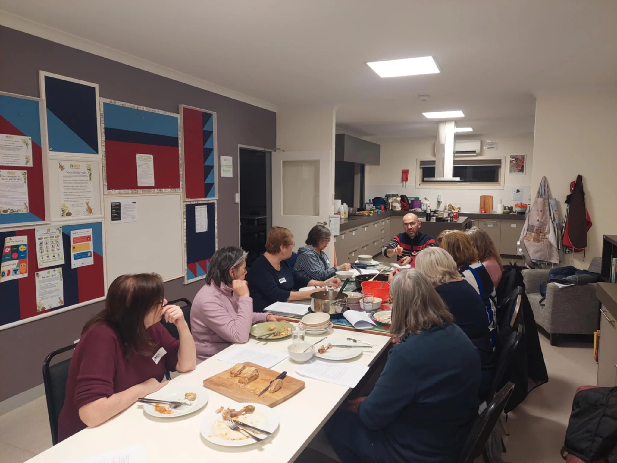Group of people sitting around a dining table, eating while the nutritionist at Vitalia Nutrition 3340 is explaining the propriety of food, in a room with modern decor and kitchen in the background.