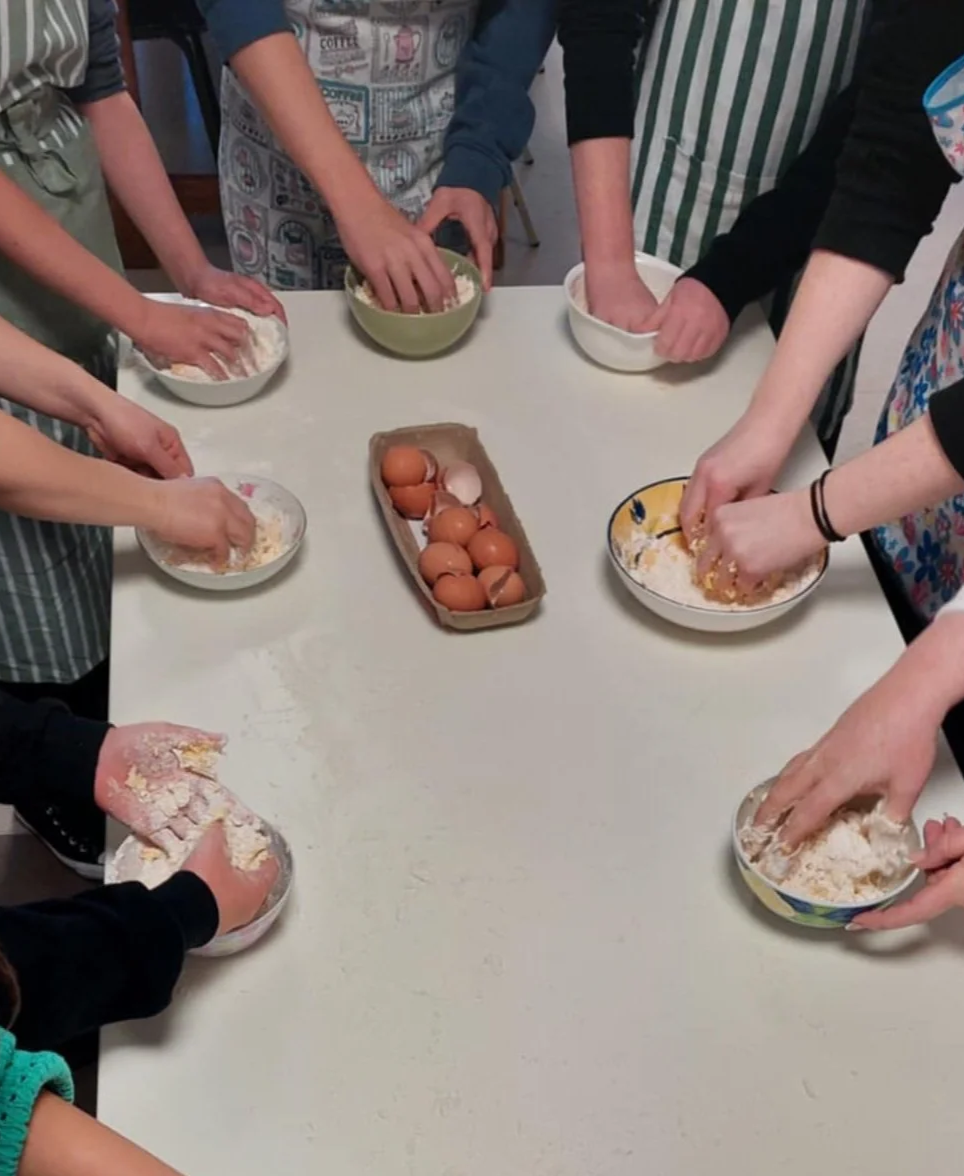 People gathered around a table, mixing ingredients by hand to prepare dough, with a tray of eggs at the centre during a workshop