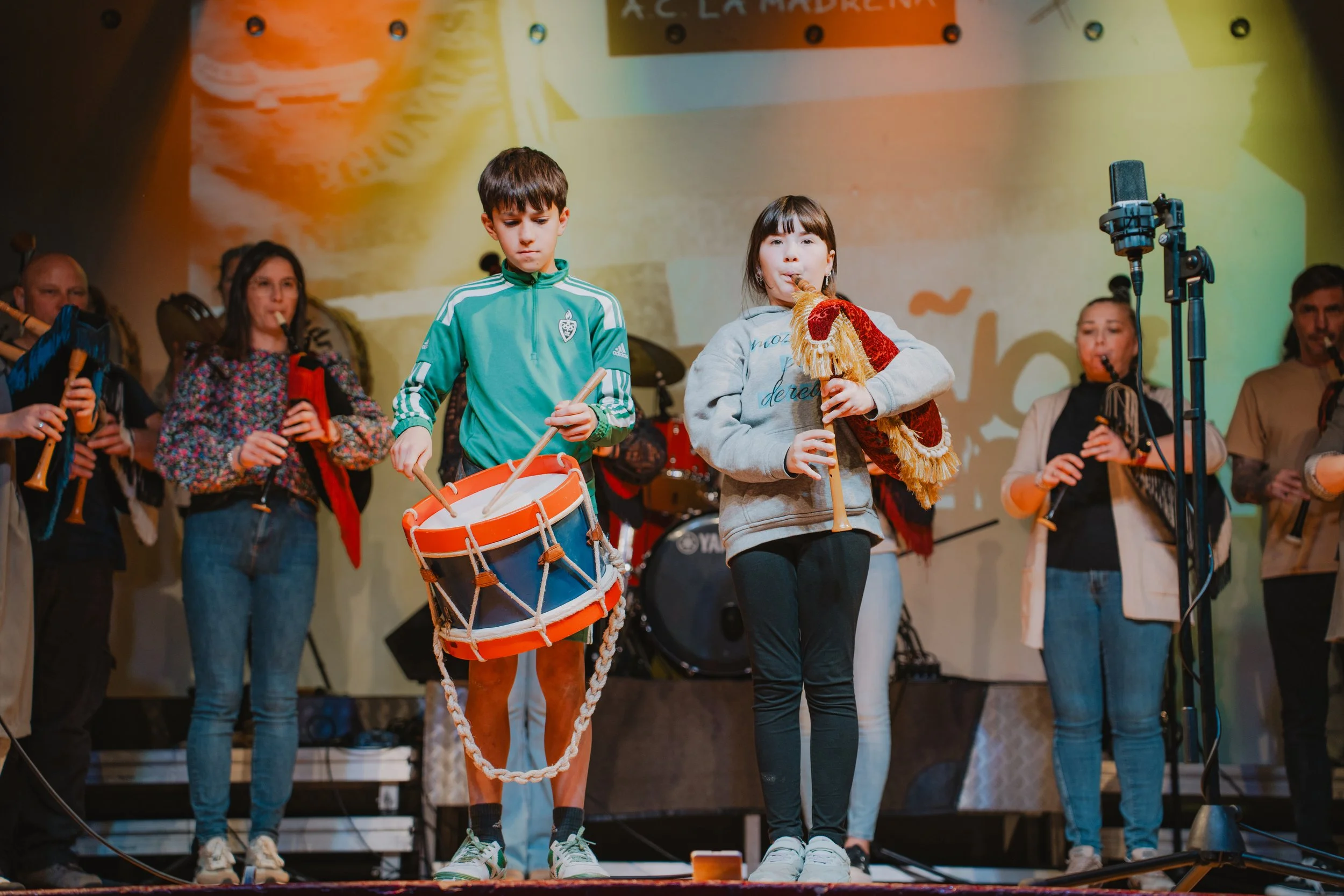 Un grupo de personas toca instrumentos musicales en un escenario. Un niño toca un tambor y una niña toca una gaita. Detrás, otras personas tocan instrumentos de viento. El fondo tiene luces y un cartel parcialmente visible. En Asturias.
