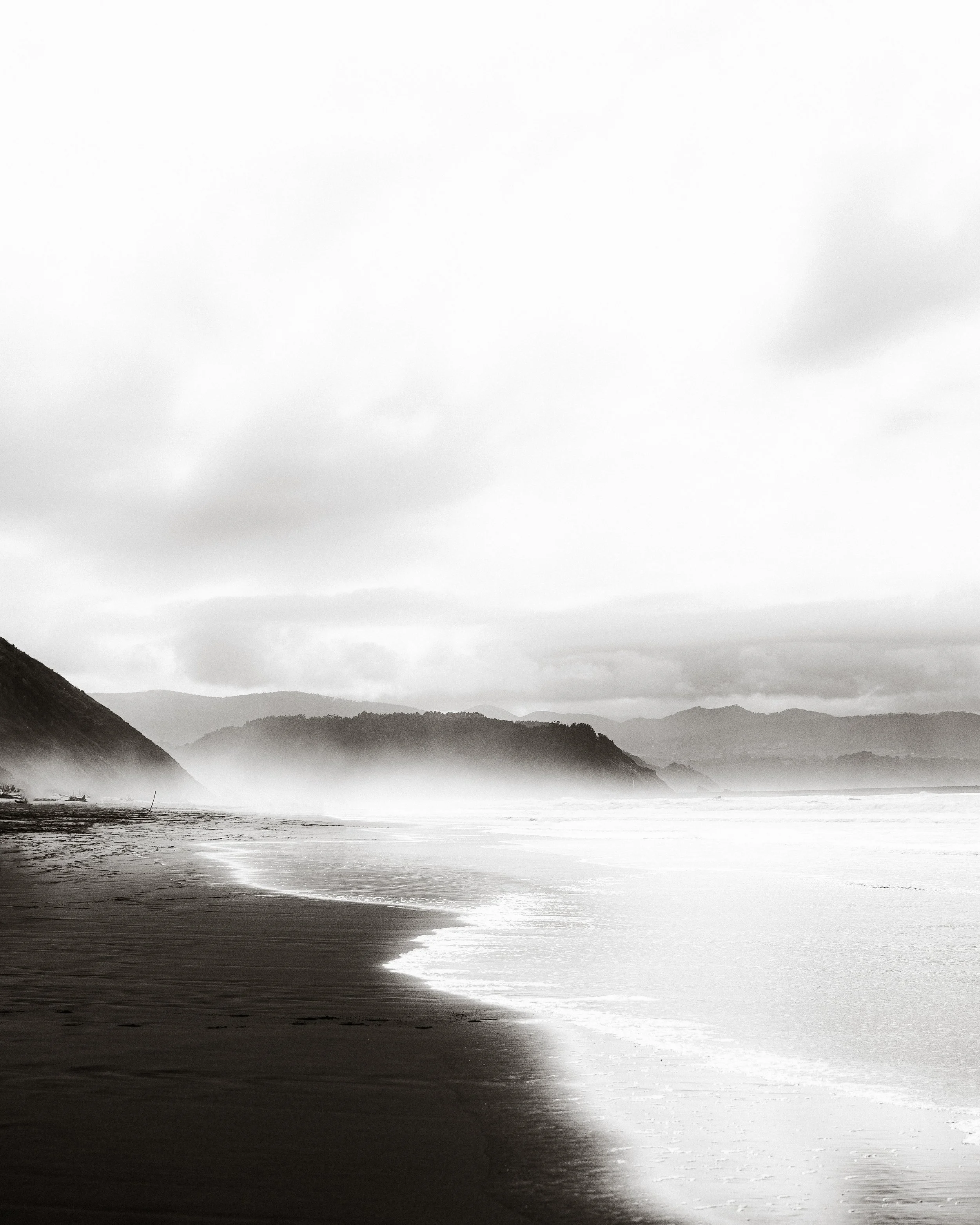Playa desierta en blanco y negro con olas suaves, cielo nublado y colinas al fondo.