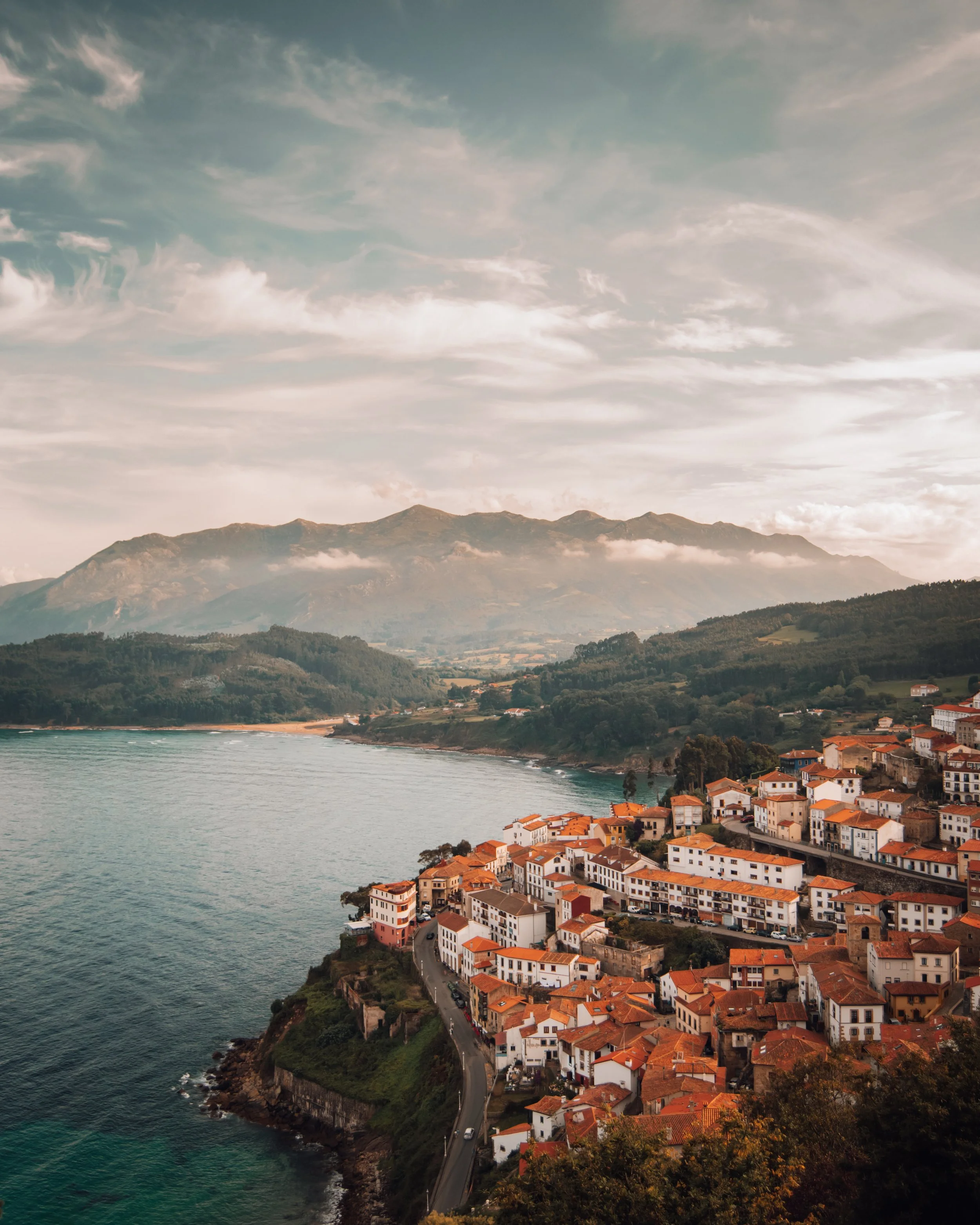 Vista panorámica de un pueblo costero de Lastres con casas de techos rojos junto al mar, rodeado de montañas y vegetación.