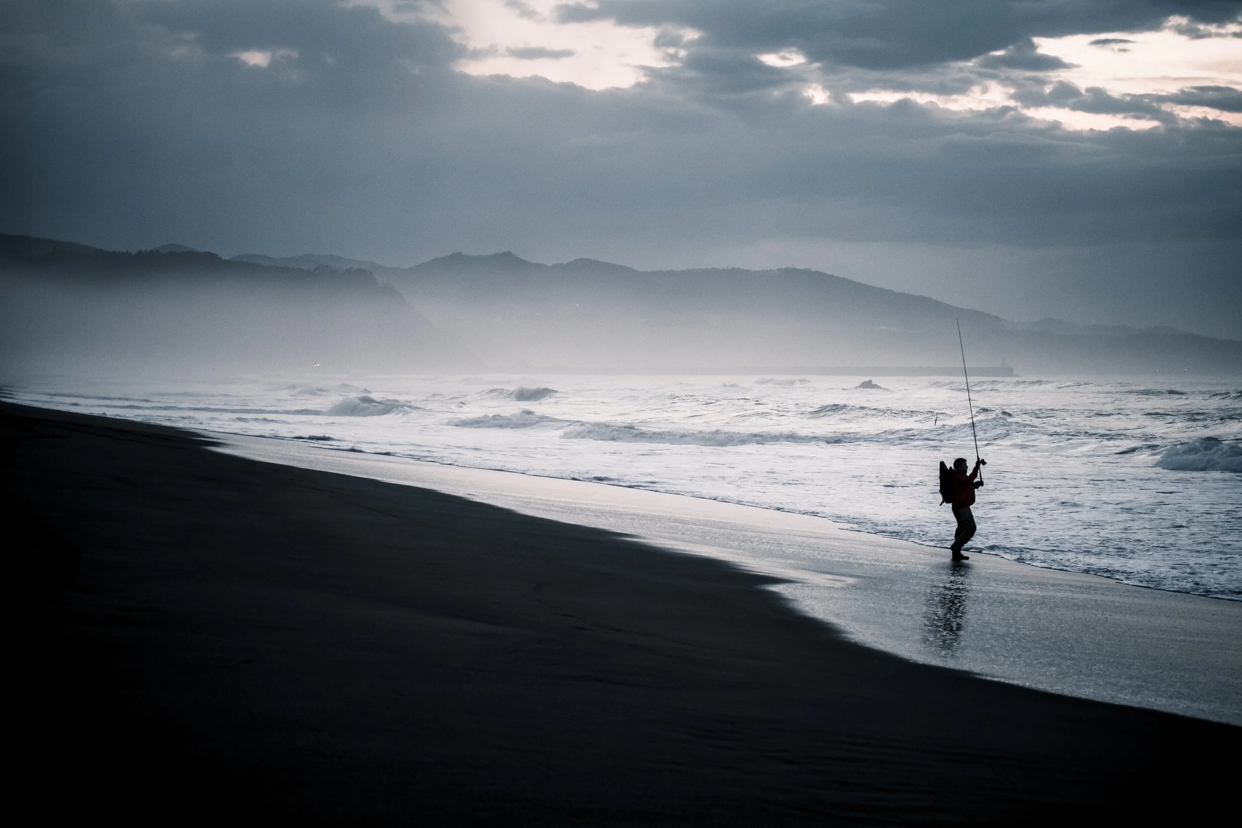 Persona pescando en la orilla de la playa al atardecer con cielo nublado y montañas al fondo.